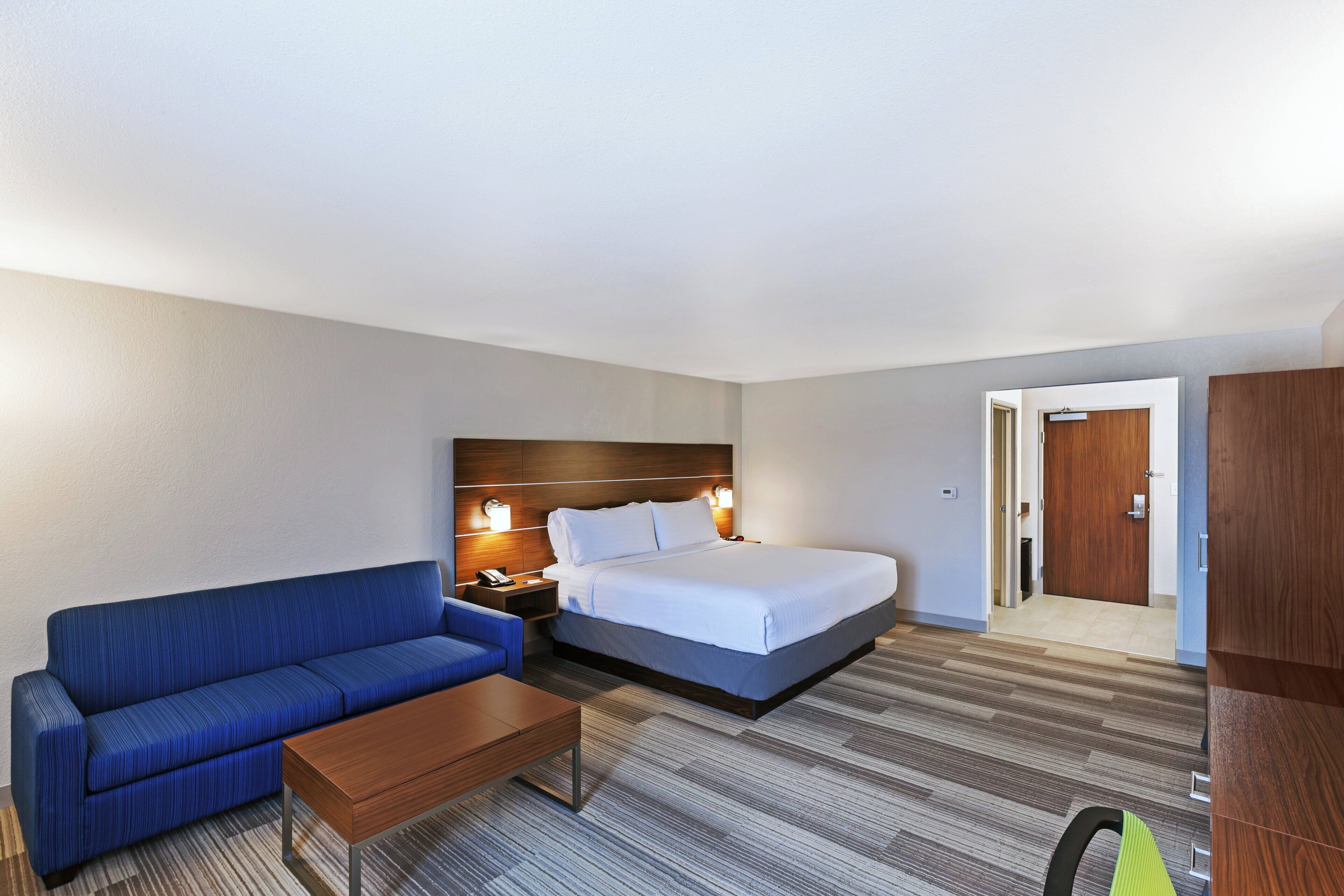 A hotel room with a bed, a blue sofa, a wooden nightstand, and a desk.