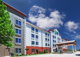 A multi-story hotel with a prominent green and white logo, a covered outdoor area, and a parking lot.