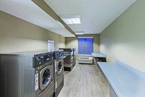 A laundry facility with multiple washing machines and dryers, a sign indicating guest laundry services, and a window with blinds.