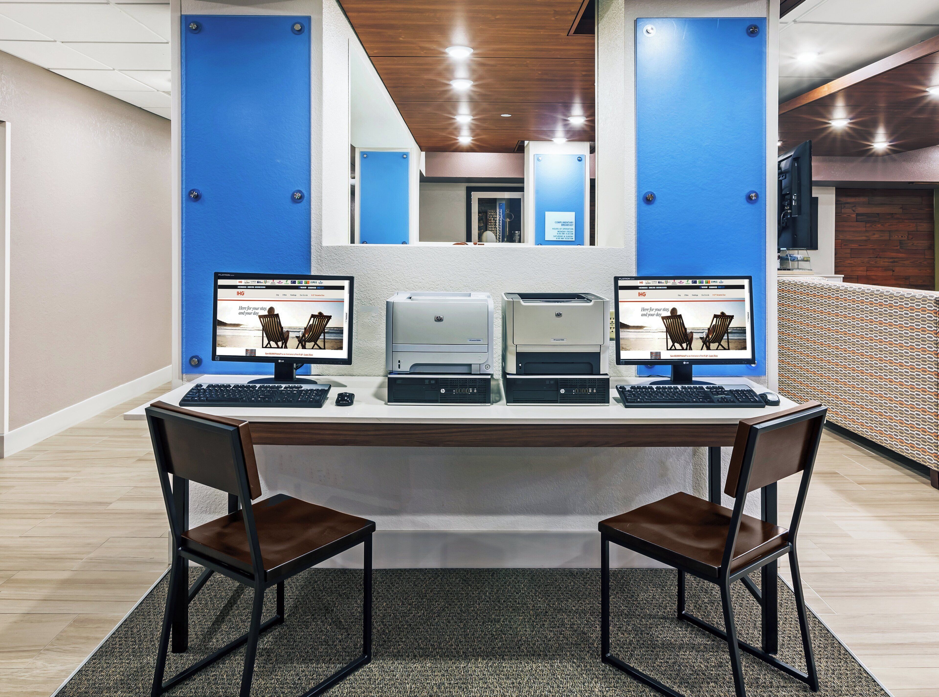 A modern office space with two desks, each with a computer monitor, keyboard, and mouse. There are two chairs in front of the desks. The room has a wooden ceiling and walls with blue accents.