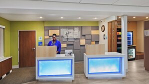 A reception area with two reception desks, a clock, and a refrigerator stocked with beverages. - Holiday Inn Express Hotel & Suites Dallas Lewisville by IHG (Lewisville)
