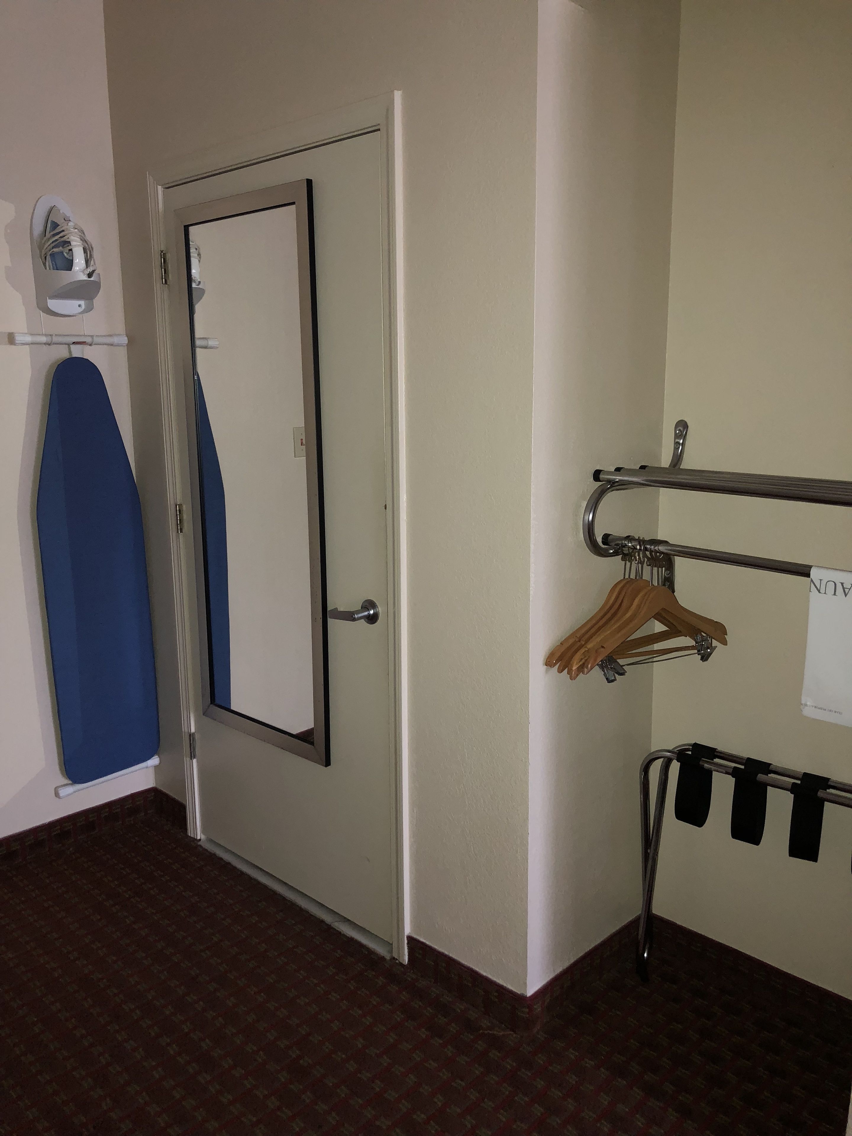 Una habitació d'hotel amb la porta tancada, una peça de roba blava penjada, una pissarra blanca i una prestatgeria metàl·lica amb penjalls.