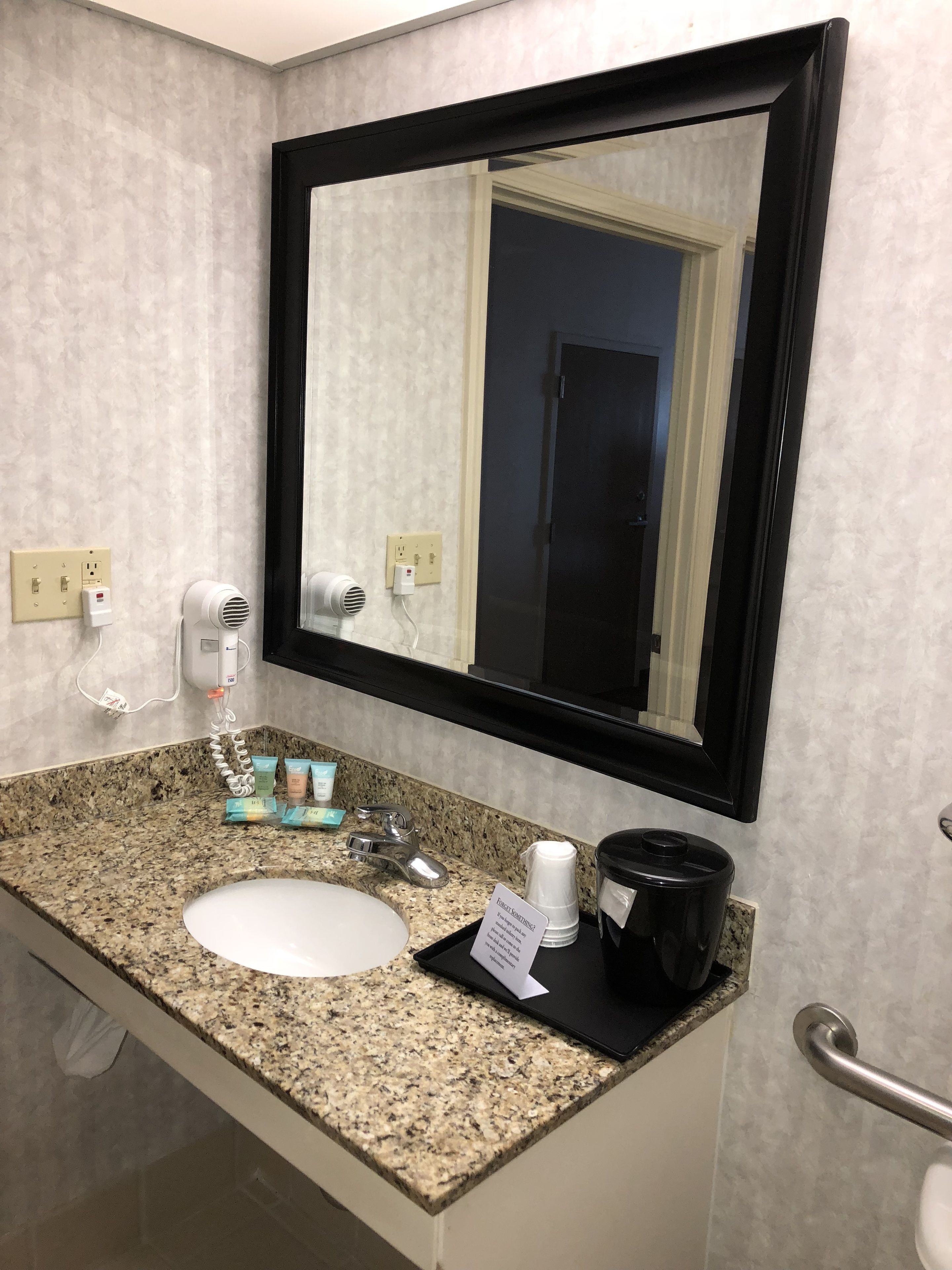 Un baño con encimera de granito, lavamanos, espejo, secador de pelo y cafetera negra.