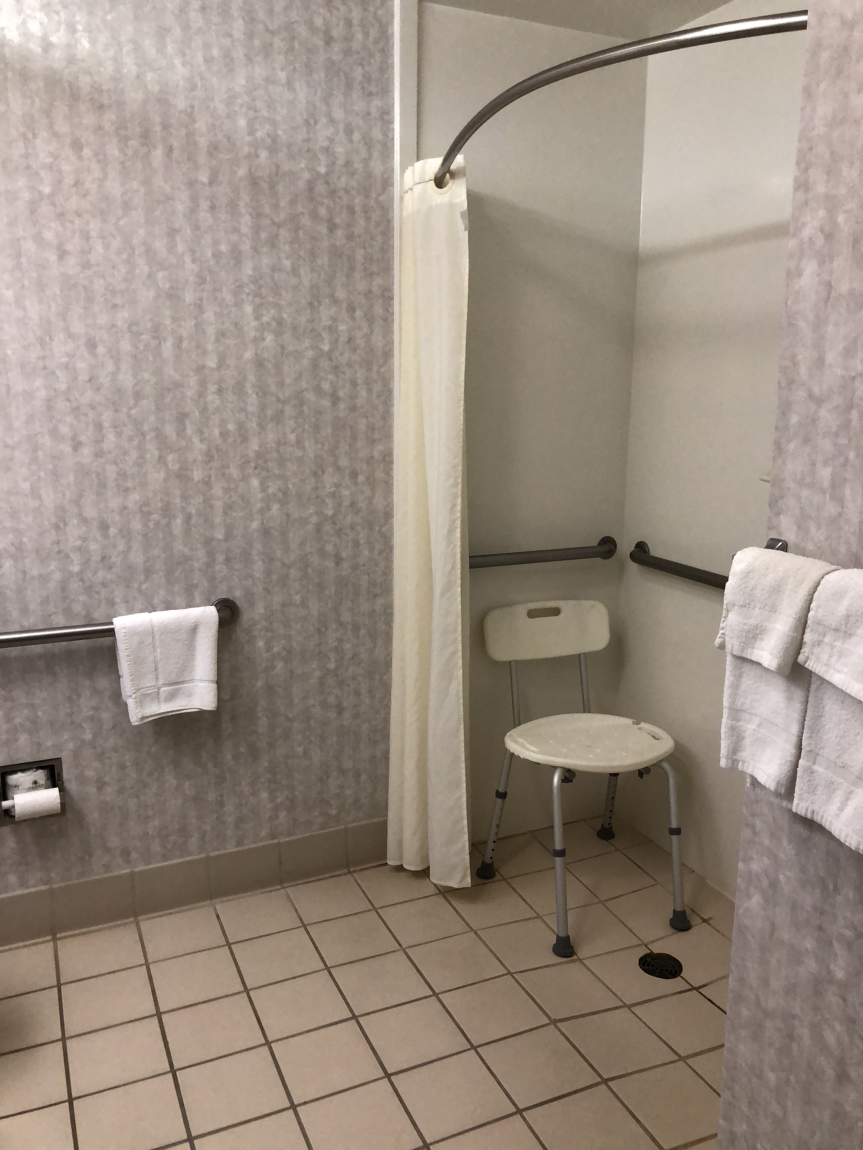 A shower area with a white shower curtain, a grab bar, a white chair, and a towel rack with towels.