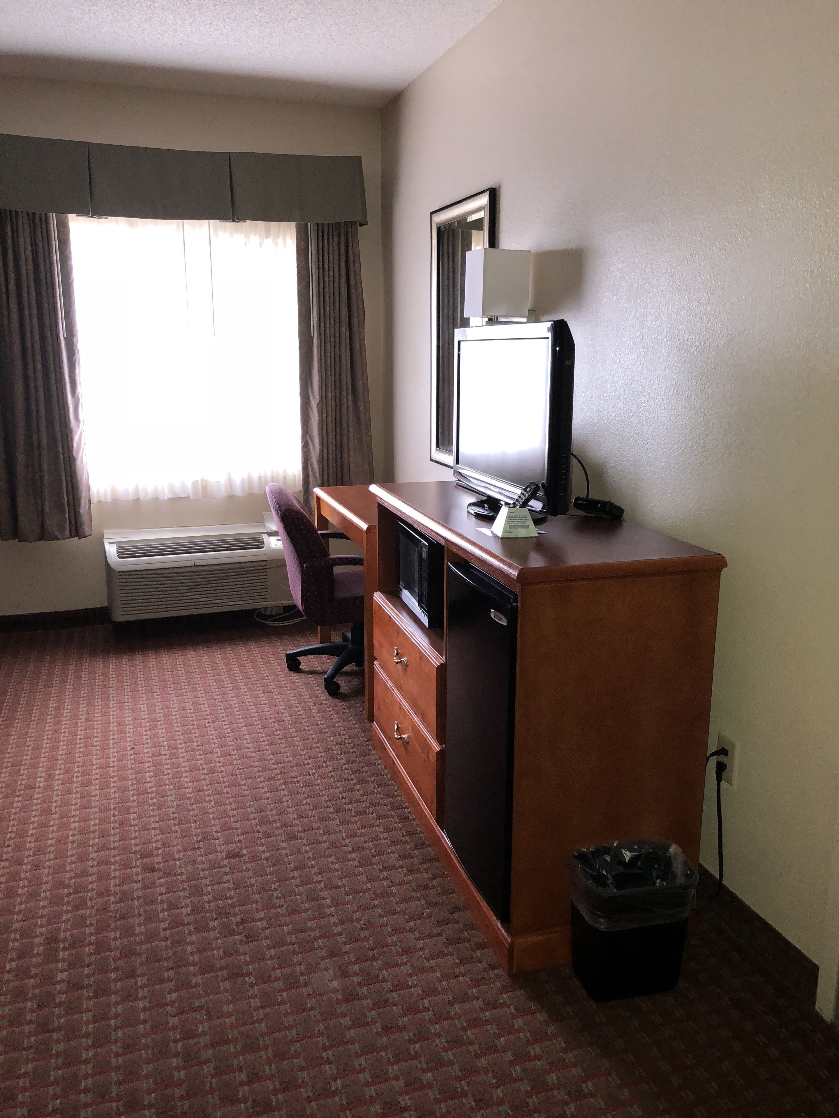 A hotel room with a desk, chair, television, and a waste bin.