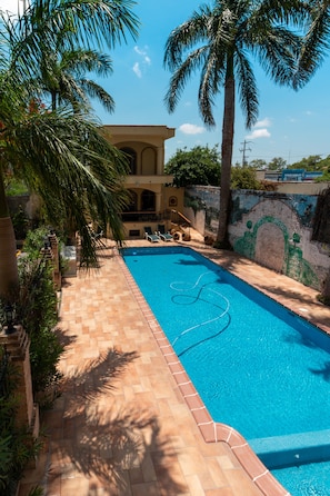Una piscina con solárium de azulejos, rodeada de palmeras y un edificio con fachada de cerámica.