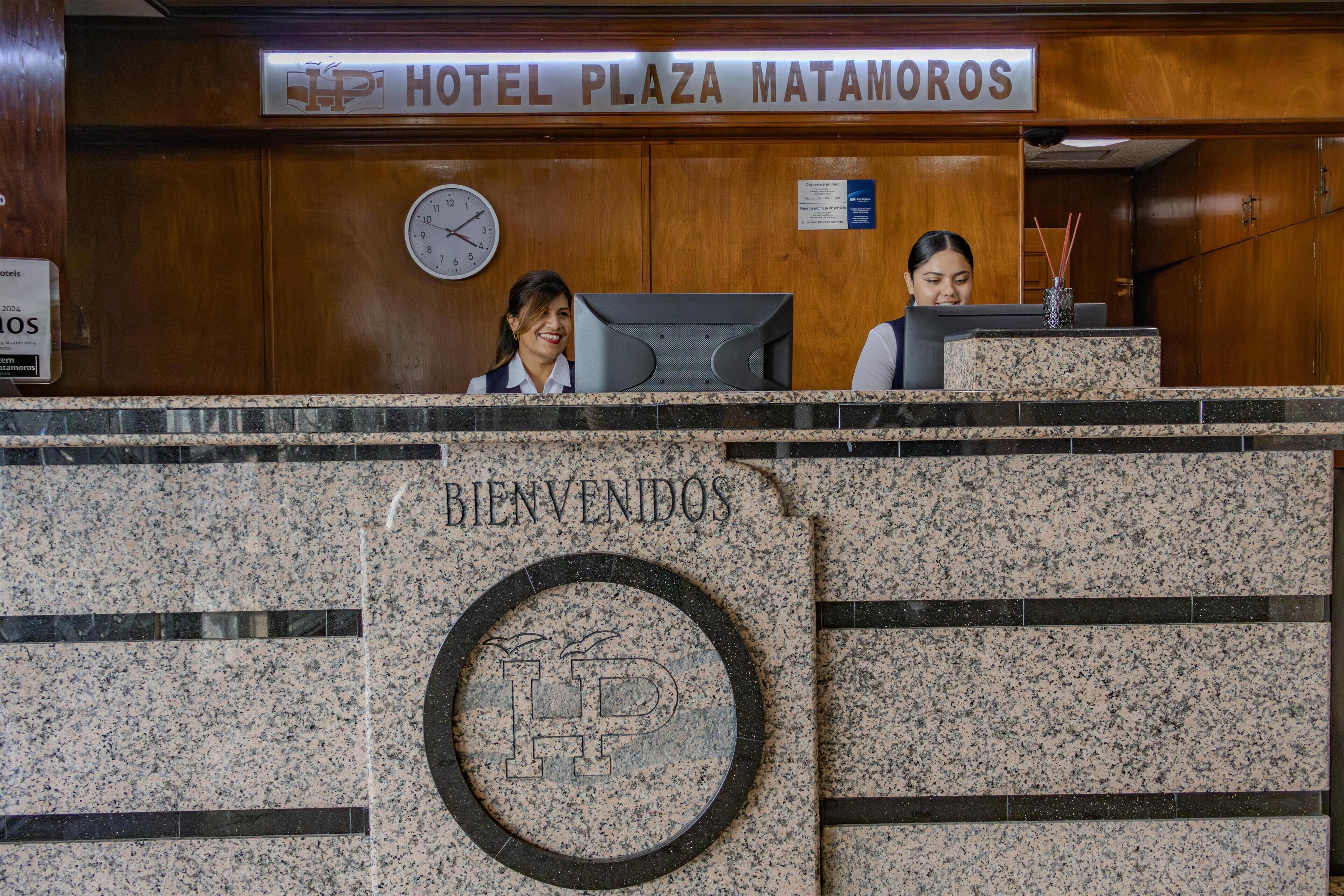 Un comptoir d’accueil à l’hôtel Plaza Matamoros, avec une horloge, une pancarte indiquant « BIENVENIDOS » et deux réceptionnistes derrière le comptoir.