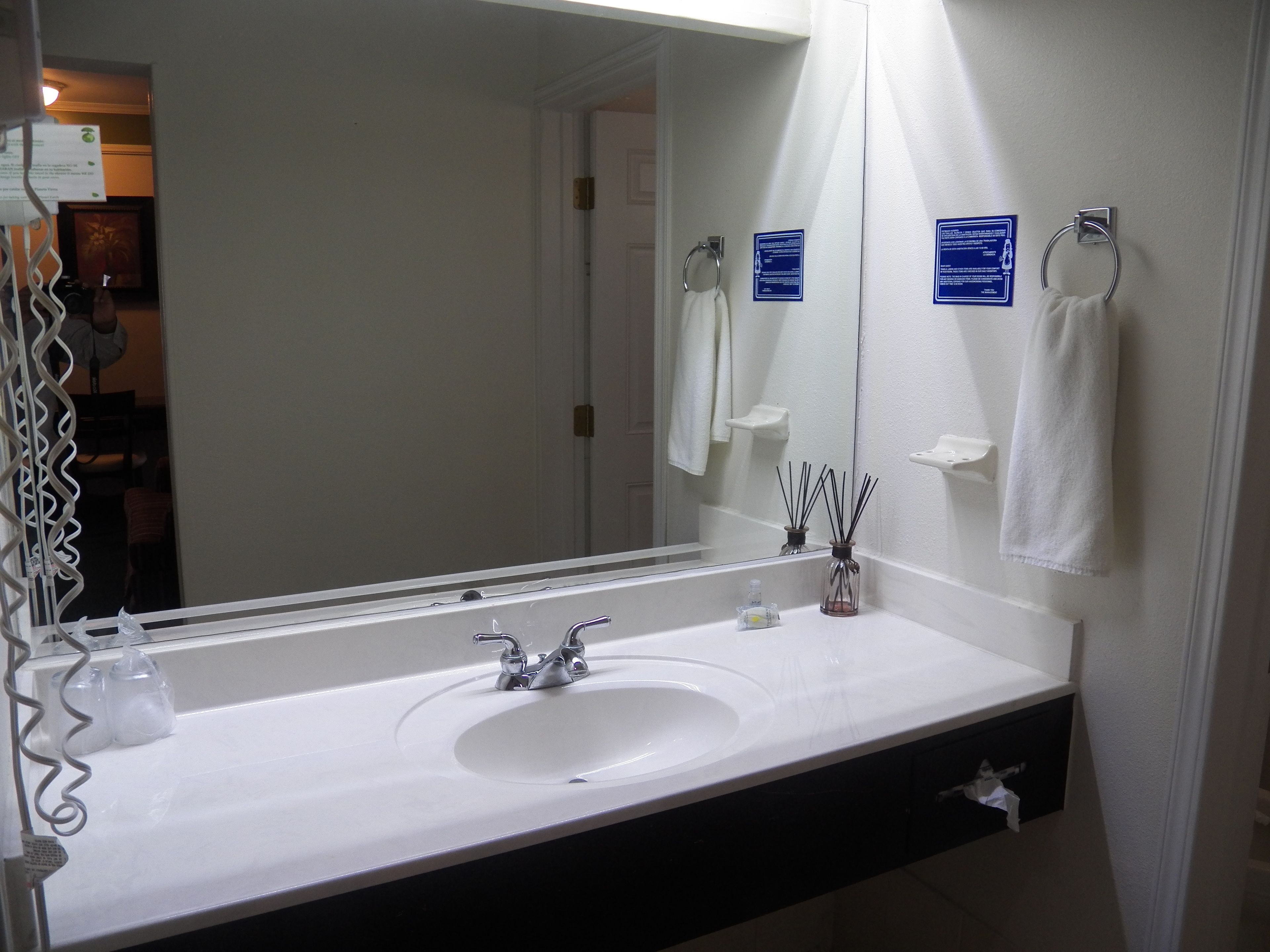 Une salle de bain avec un lavabo blanc, un grand miroir et deux panneaux d’information bleus accrochés au mur.