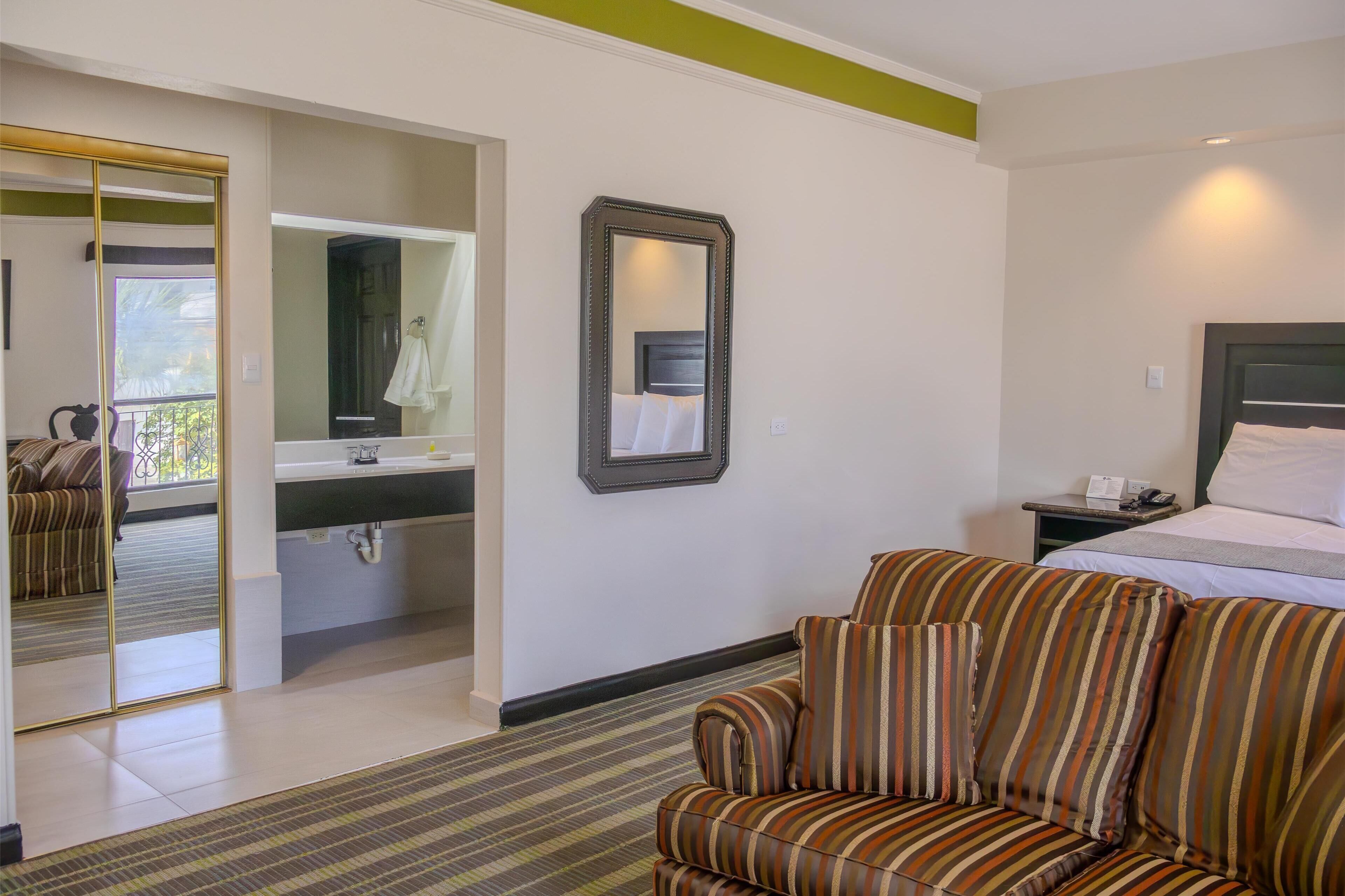 A hotel room with a bed, a striped sofa, a mirror, and a bathroom visible through an open door.