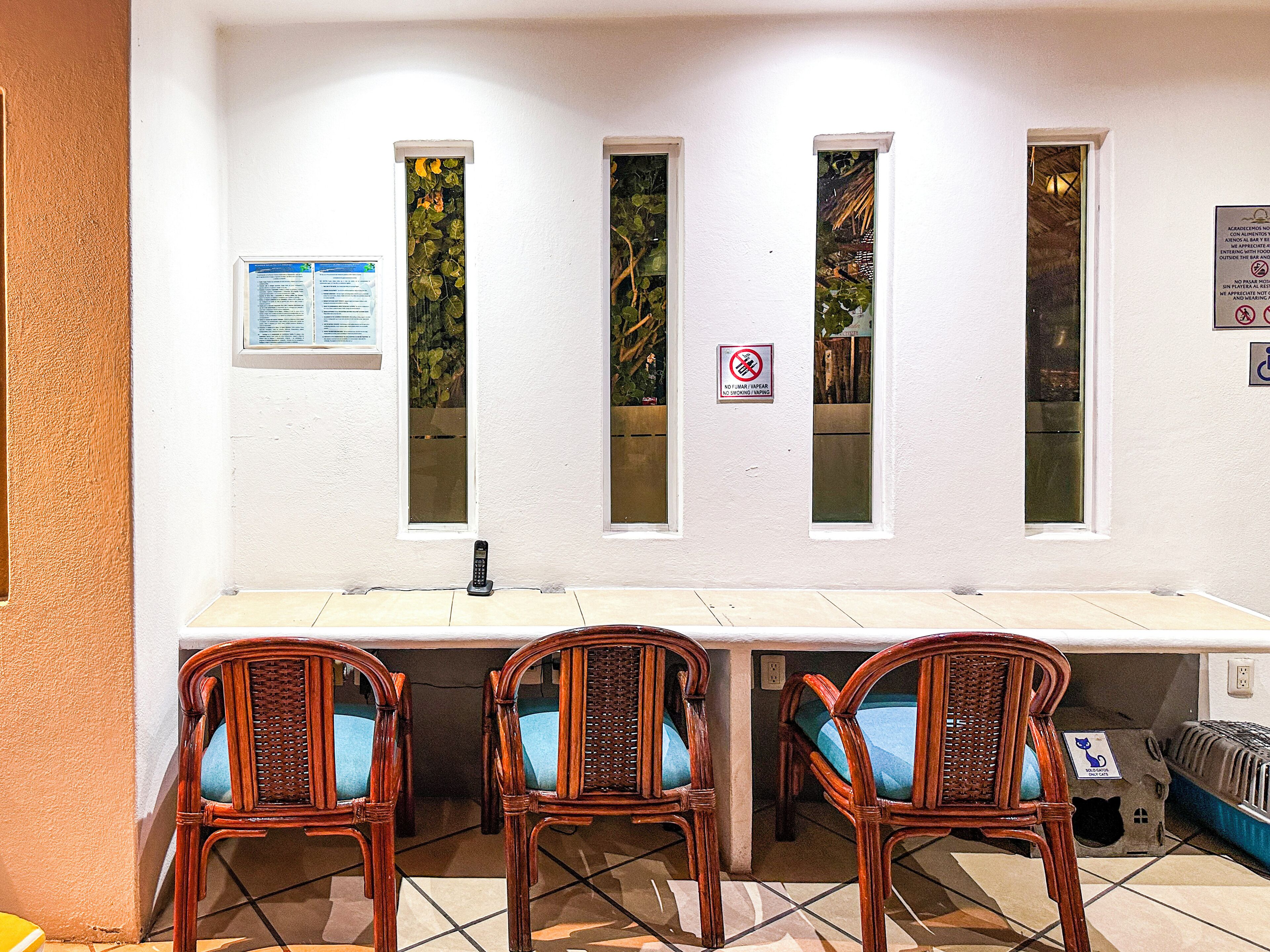 A room with a white wall, three windows with plants, a no smoking sign, and a table with chairs.
