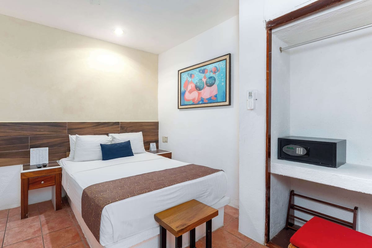 Una camera d'albergo con una porta in legno, un letto con biancheria da letto bianco e blu, un comodino con un telecomando, un quadro appeso al muro, una cassaforte nera e una sedia rossa.