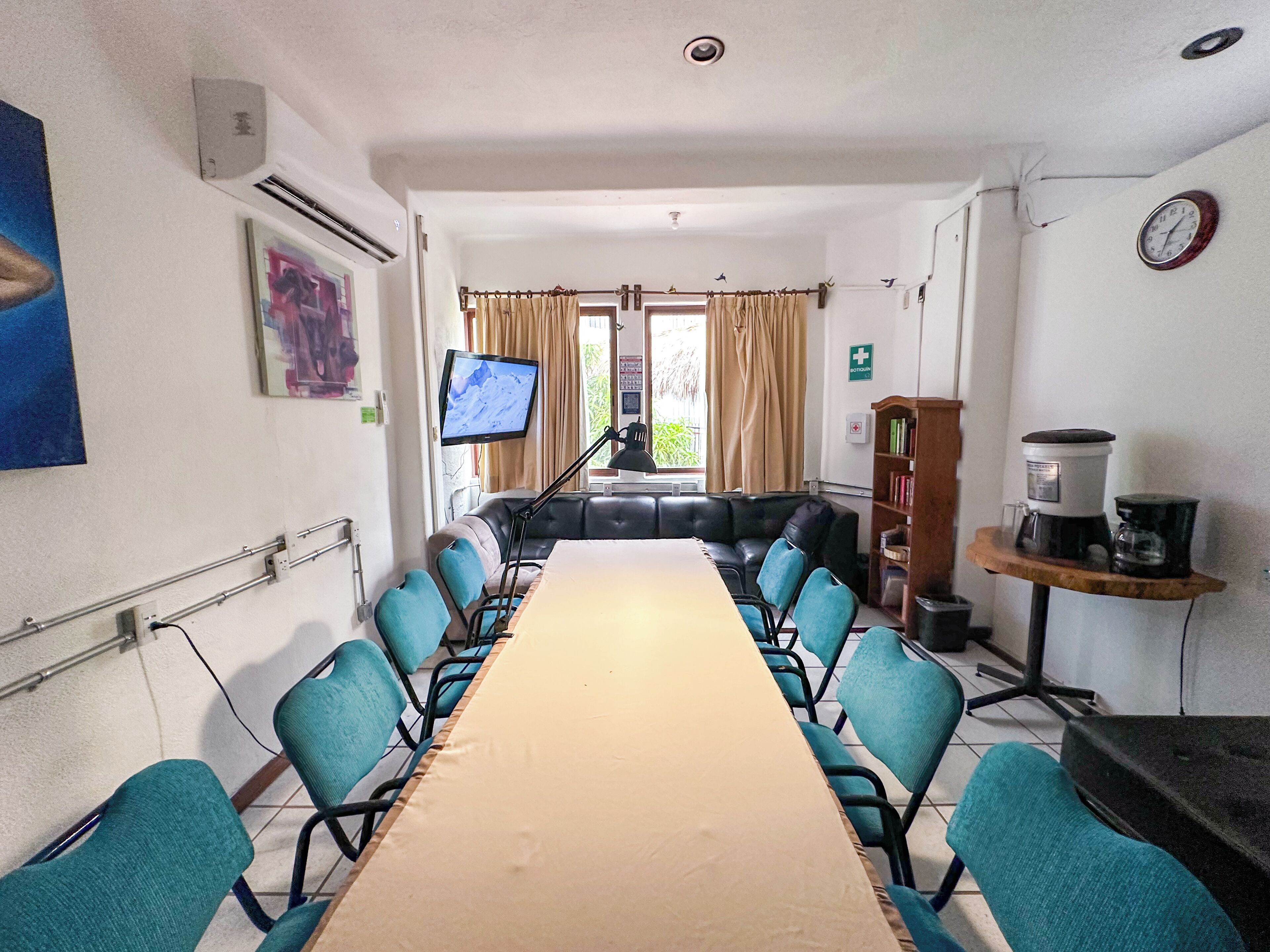 A conference room with a long table, blue chairs, a television, a bookshelf, and a clock on the wall.