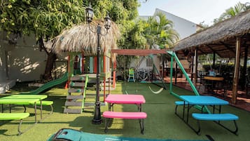 Explore the outdoor play area with a slide, swings, and climbing frame, surrounded by lush greenery and palm trees.