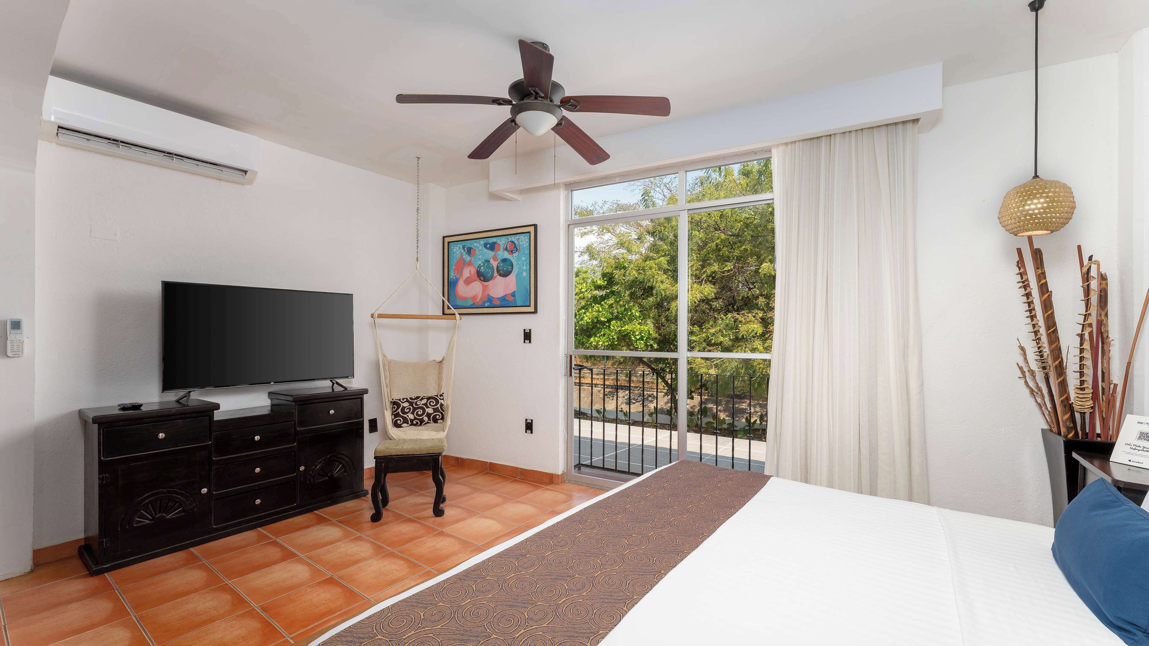 A hotel room with a flat-screen TV, a ceiling fan, a large window with a view of greenery, a hanging chair, and a bed with a white headboard.