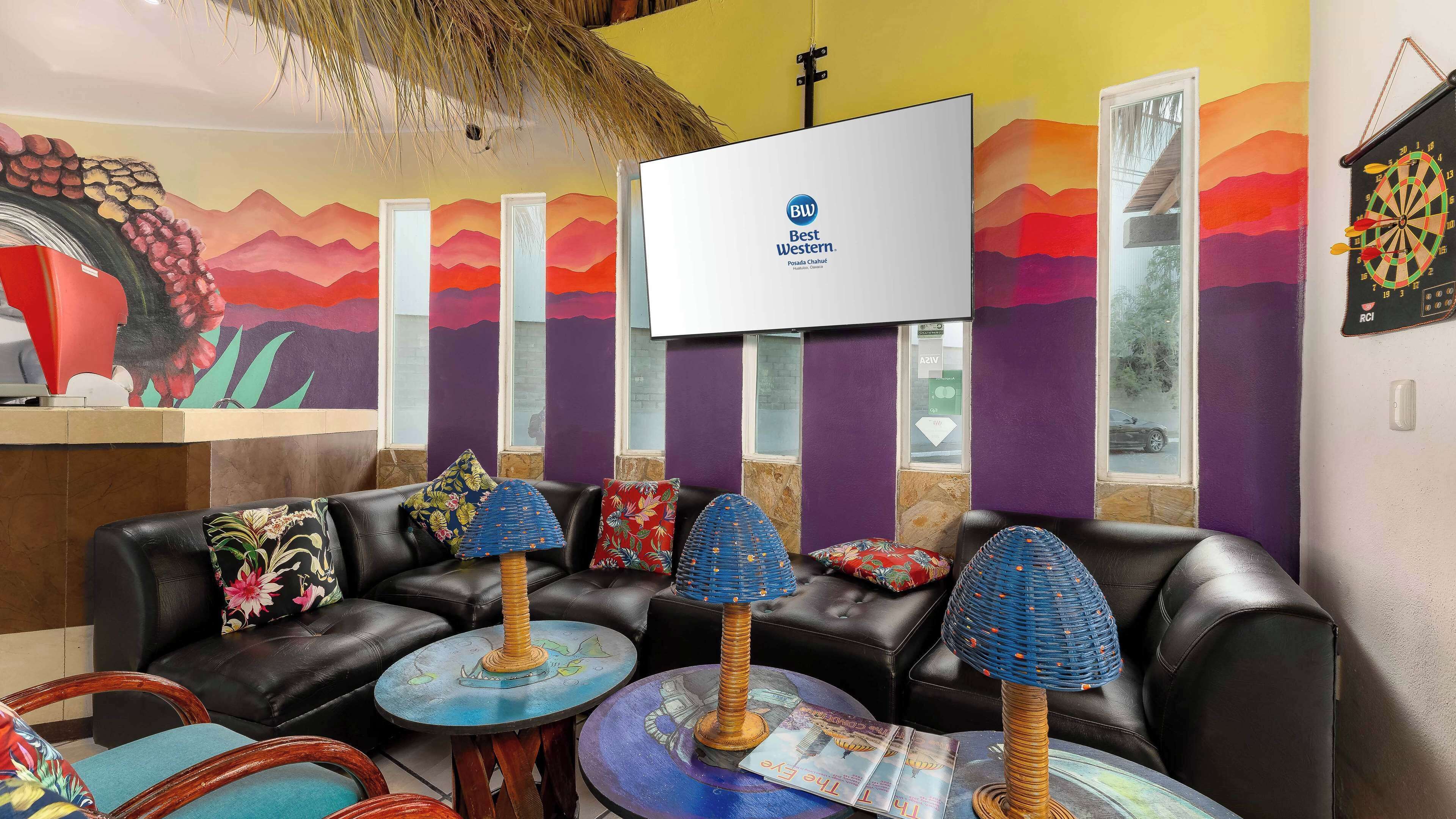 A modern lounge area with a black leather sofa, colorful cushions, and a unique blue lamp. A large TV screen displays the logo "Best Western."