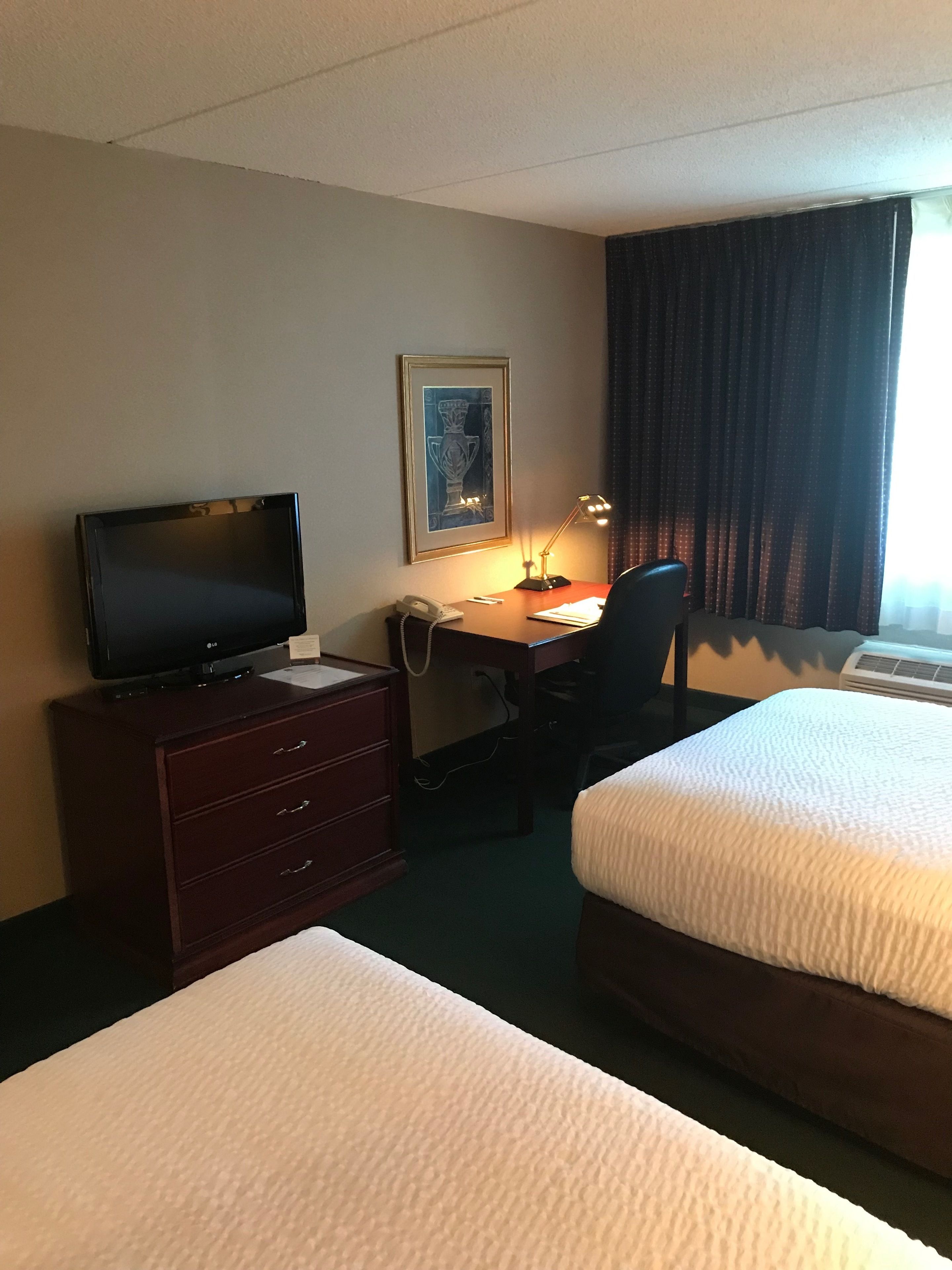 A hotel room with two beds, a desk with a computer and phone, a TV, and a framed picture on the wall.