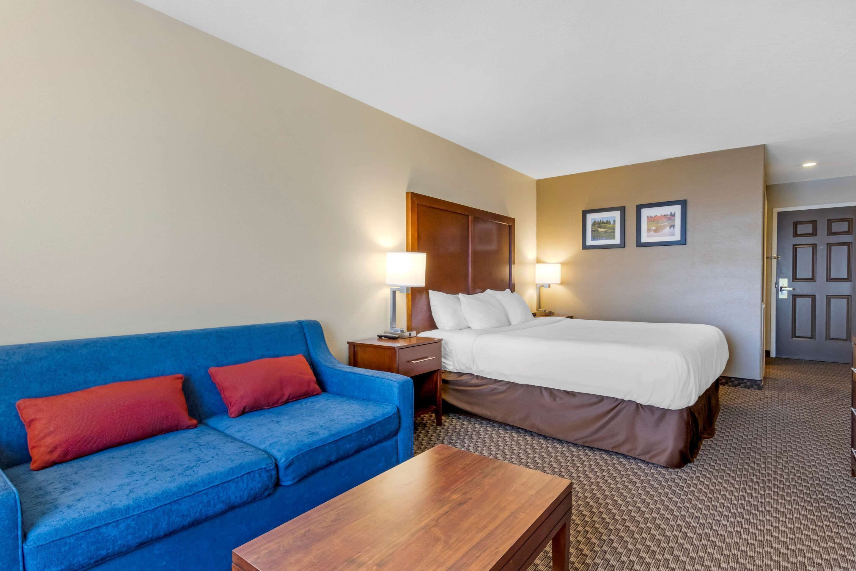 A hotel room with a blue sofa, a wooden table, a bed with white linens, two bedside lamps, and a door leading to another room.