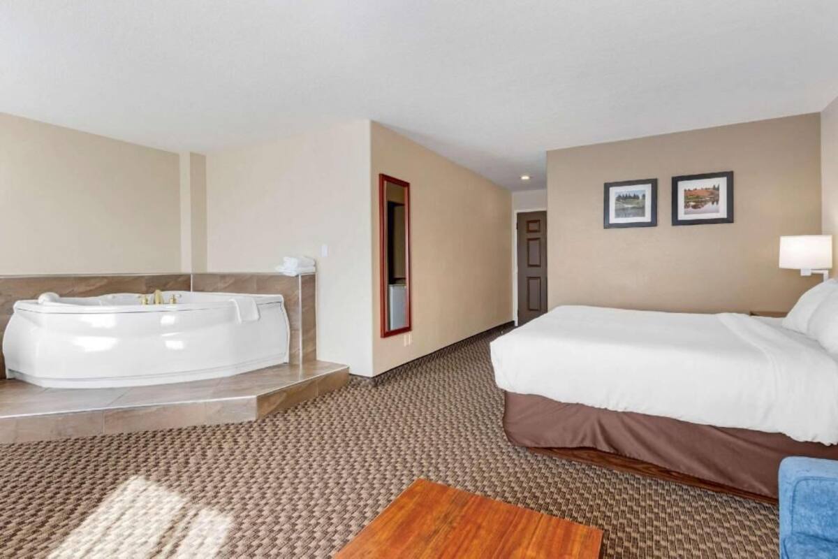 Habitación de hotel con una bañera grande, una cama, una mesita y dos cuadros en la pared.