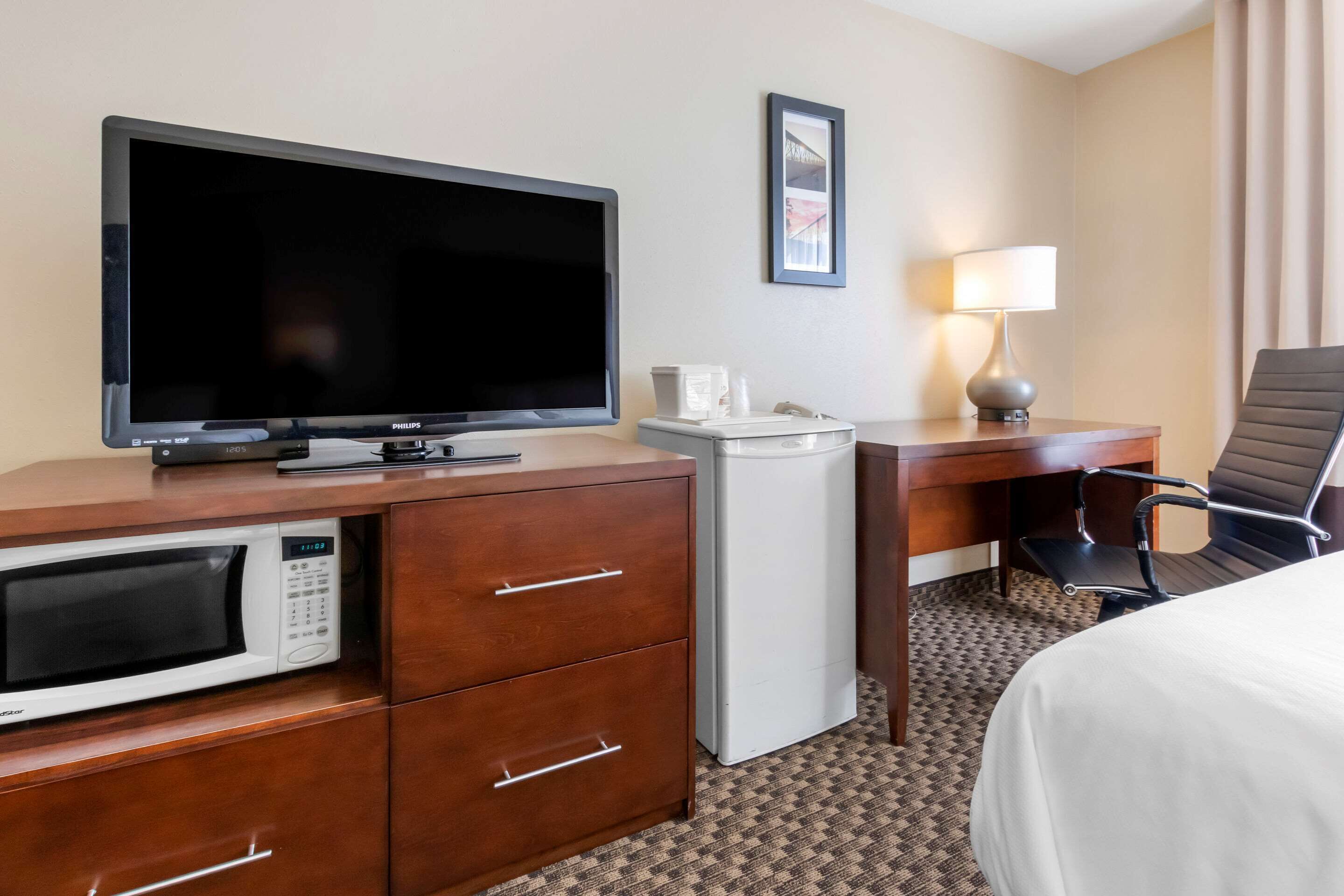Habitación de hotel con televisión de pantalla plana, microondas, refrigerador pequeño, escritorio con lámpara, una silla y una cama.