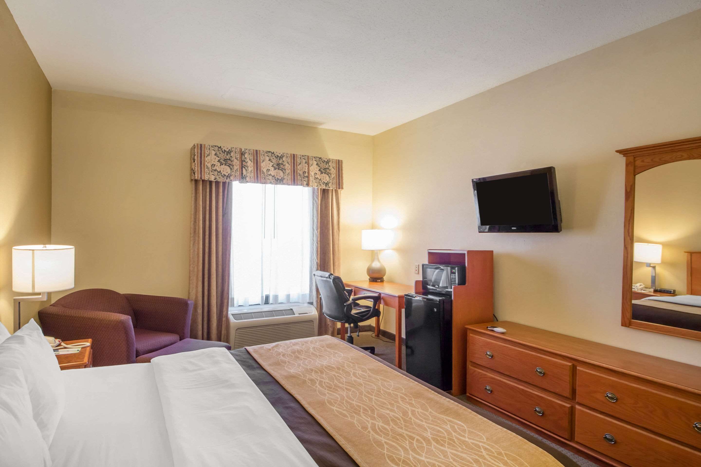 Een hotelkamer met een bed, een bureau met een computer, een televisie, een dressoir en een raam met gordijnen.