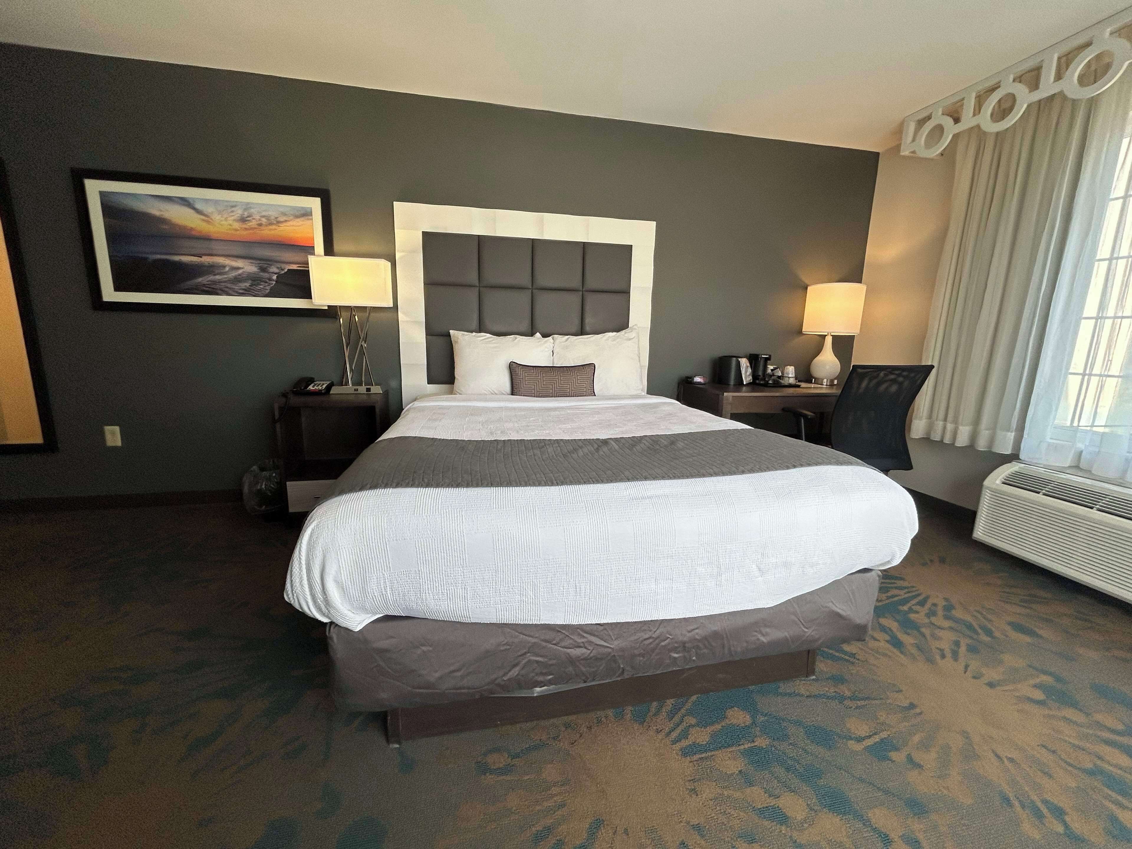 A hotel room with a large bed, a nightstand with a lamp, a desk with a chair, and a window with curtains.