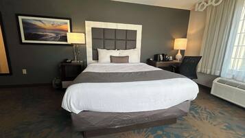 A hotel room with a large bed, a nightstand with a lamp, a desk with a chair, and a window with curtains.