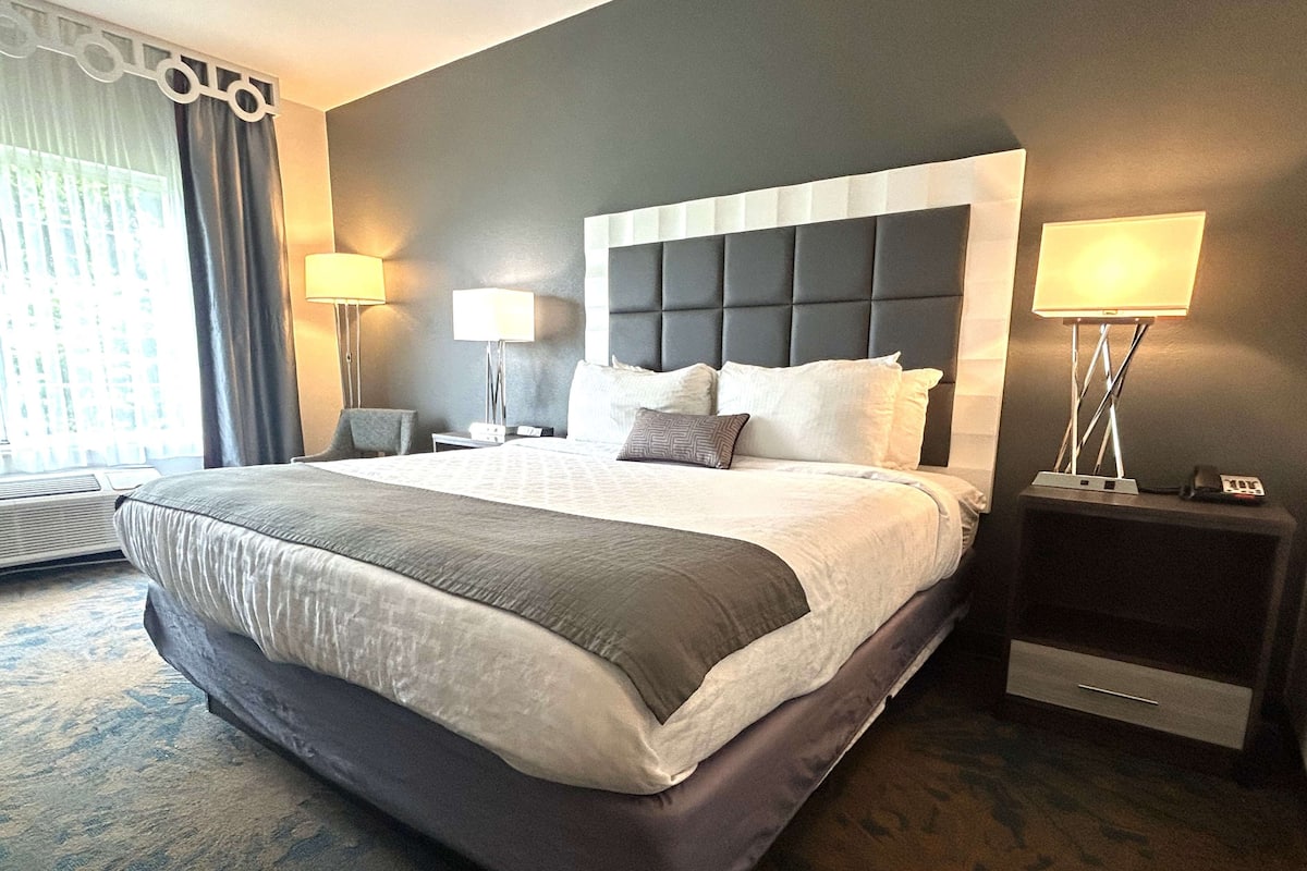 A hotel room with a large bed, two bedside tables with lamps, a window with curtains, and a wall-mounted air conditioning unit.