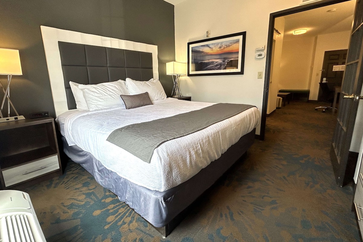 A hotel room with a large bed, a nightstand with a lamp, a framed picture on the wall, and a carpeted floor.