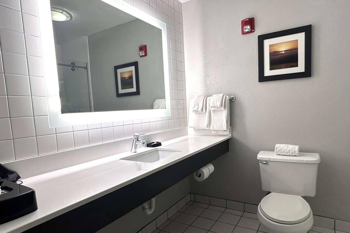 A modern bathroom with a white sink, mirror, and toilet. There are two framed pictures on the wall, a towel rack, and a fire alarm.