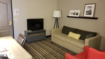 A hotel room with a flat-screen TV on a stand, a sofa, a red chair, a desk with a phone, and a lamp.