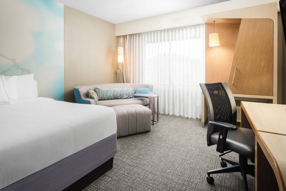 Una habitación de hotel con cama, escritorio, silla, una mesita y ventana con cortinas.