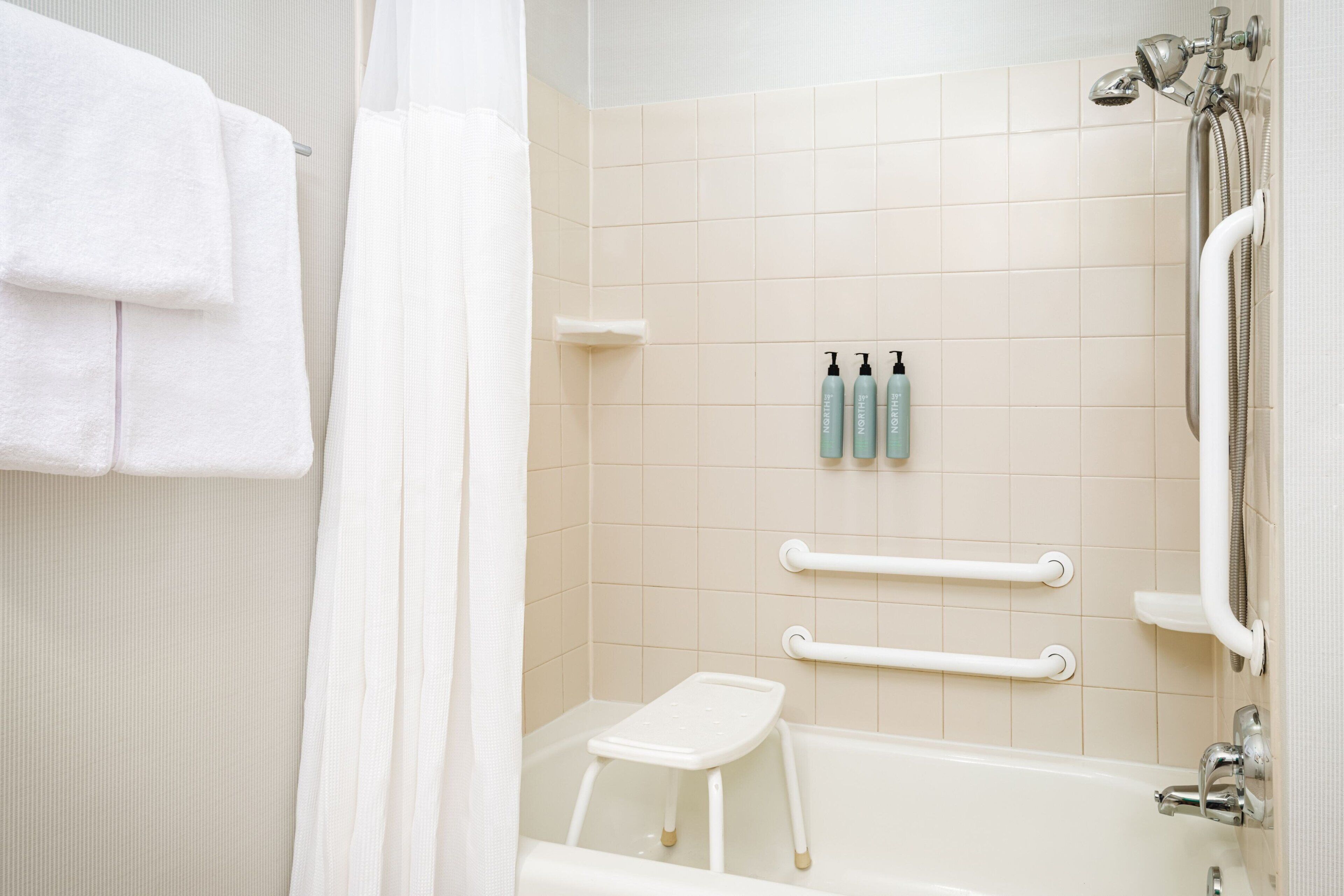 Une salle de bain avec une baignoire, une douche et un petit tabouret blanc. Deux bouteilles de shampoing et d’après-shampoing sont posées sur le mur.