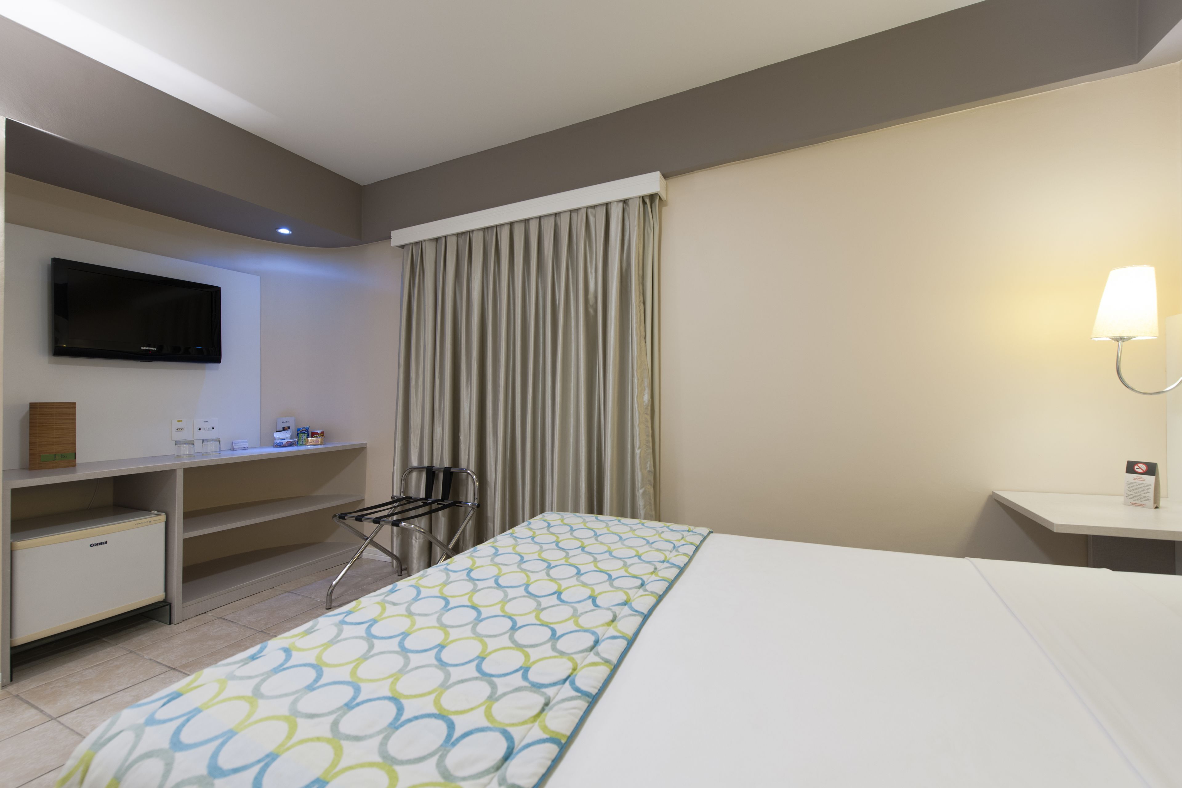 Quarto de hotel com cama, escrivaninha, televisão e banheiro.