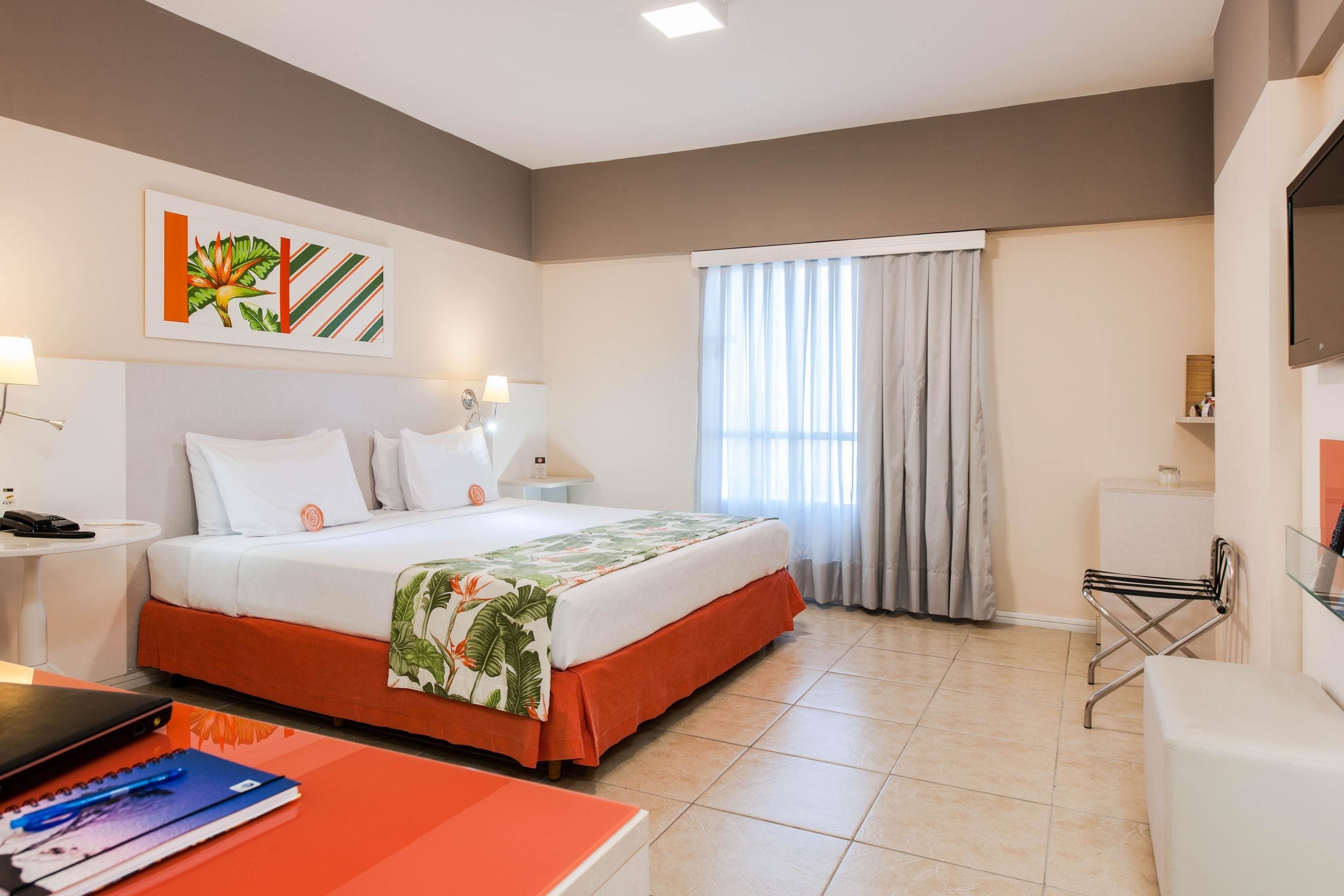 A hotel room with a bed, a desk, a chair, a TV, and a window with curtains.