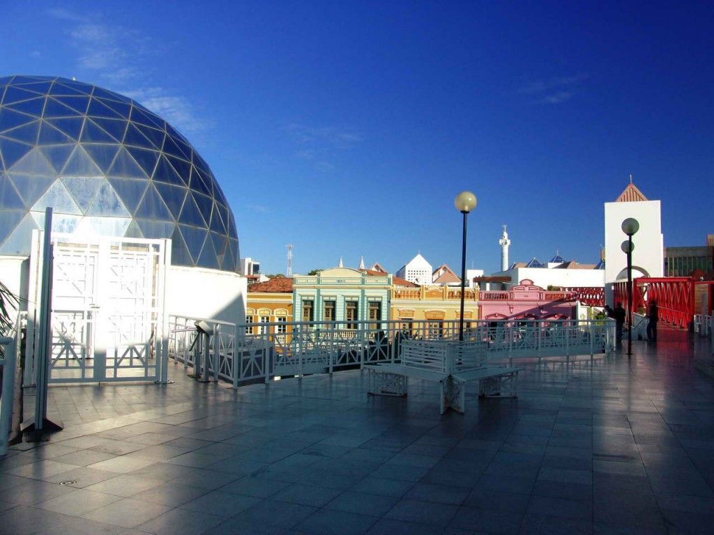 A plaza with a large glass dome structure, a white building with a red roof, and colorful buildings in the background.
