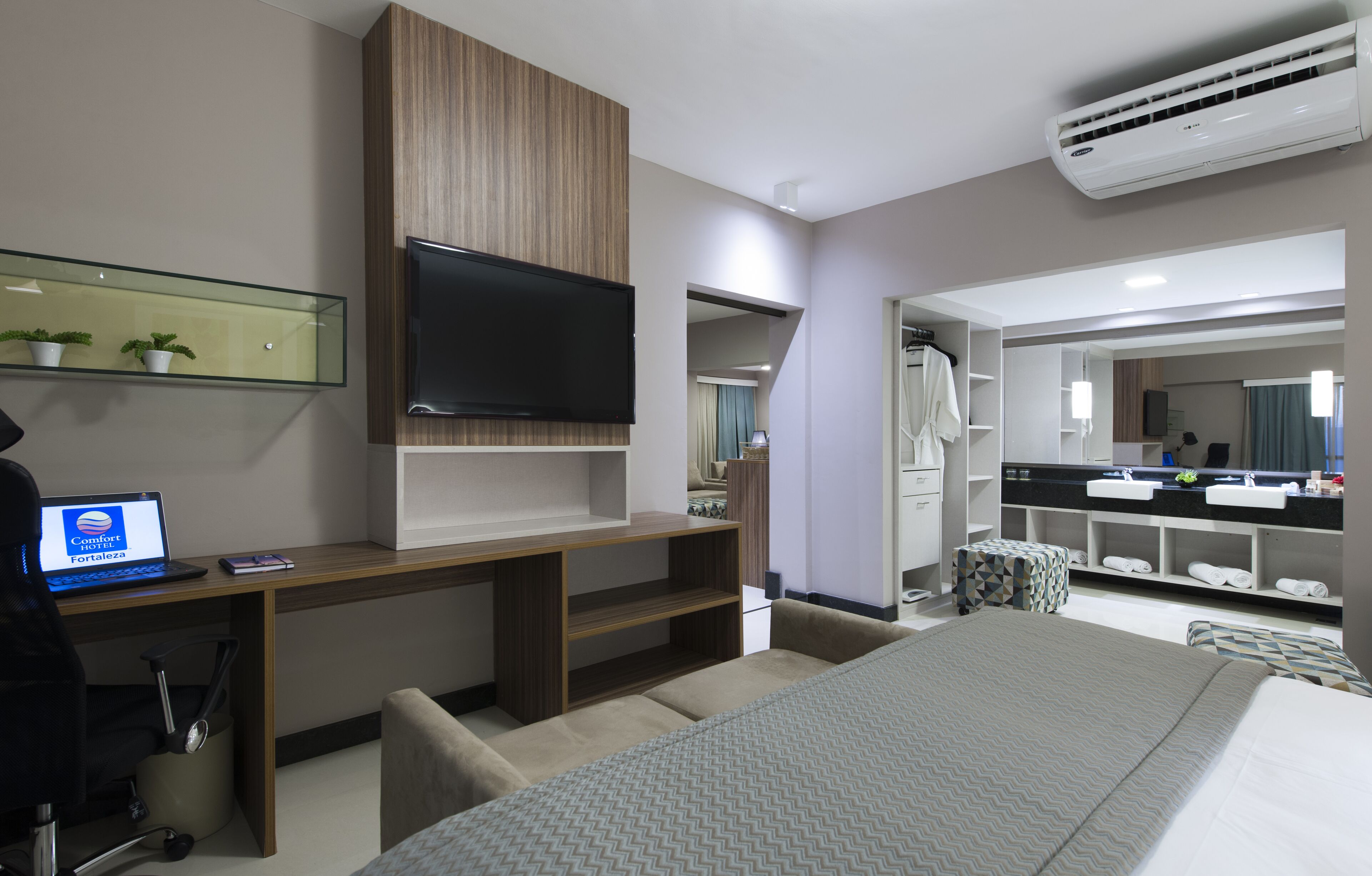 Una habitación de hotel moderna con una cama grande, un escritorio con una computadora portátil, un televisor de pantalla plana, un vestidor y un baño con tocador y espejo.
