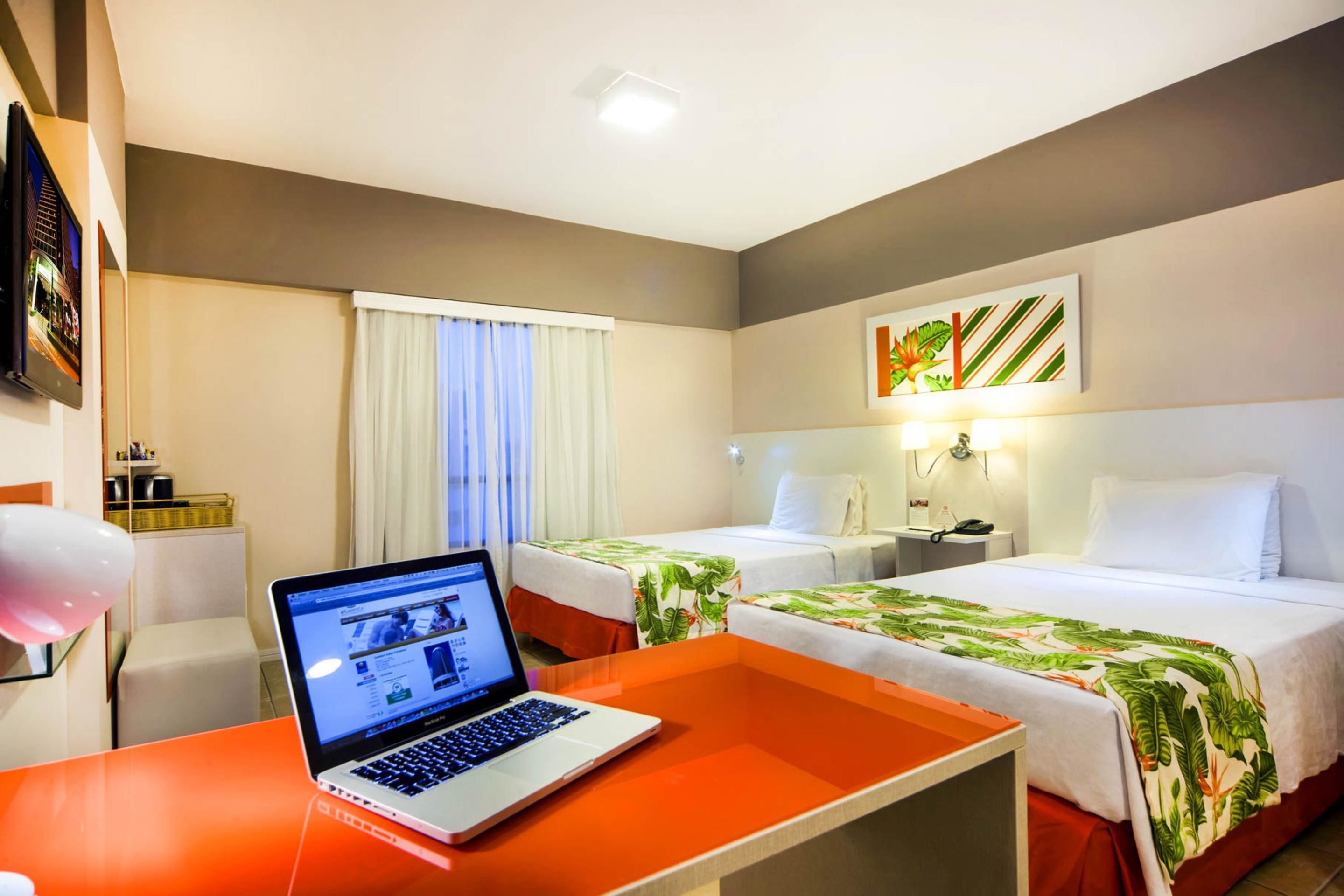A hotel room with two beds, a laptop on a table, a TV on the wall, and a painting.