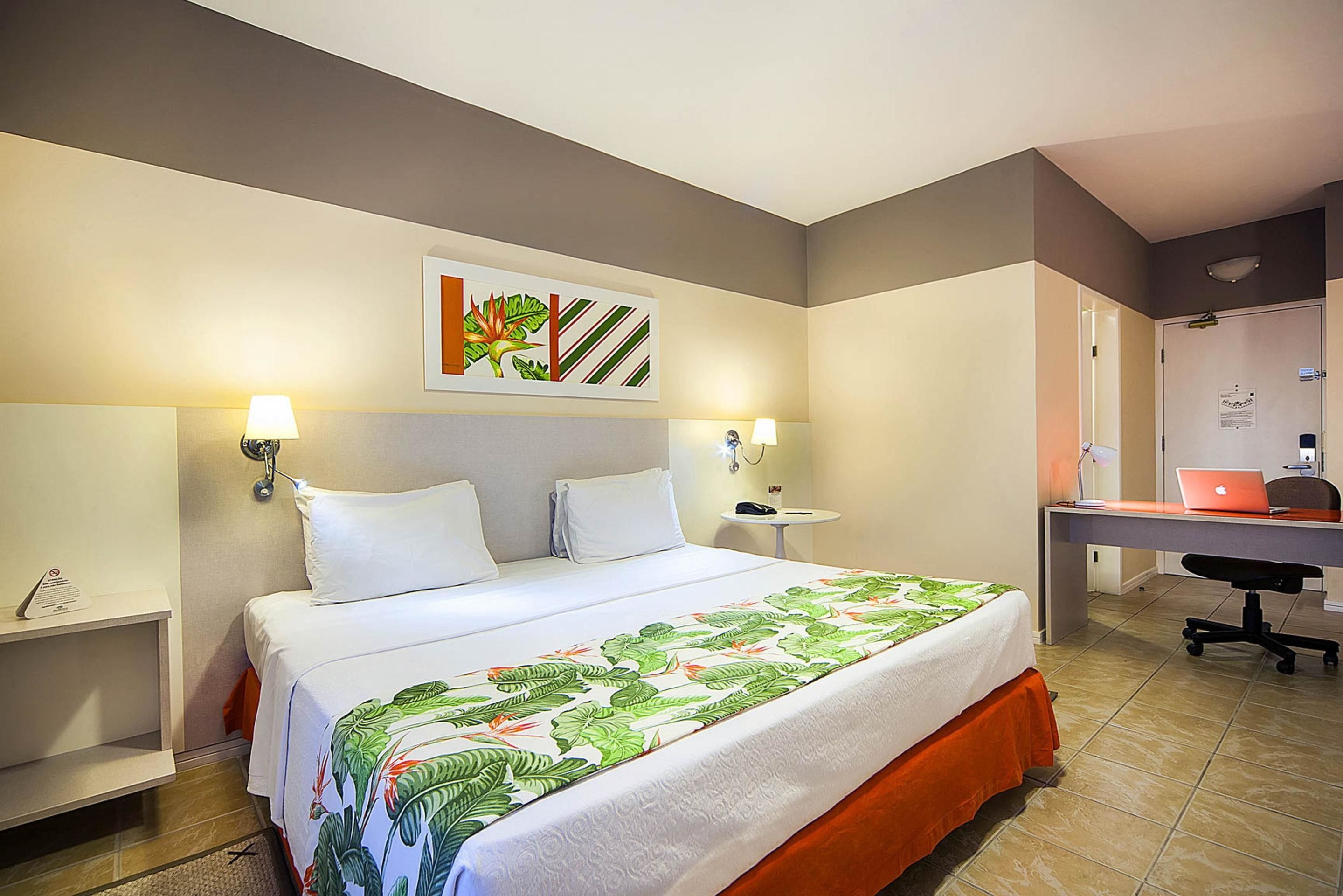A hotel room with a bed, two bedside lamps, a desk with a laptop, and a door leading to a bathroom.