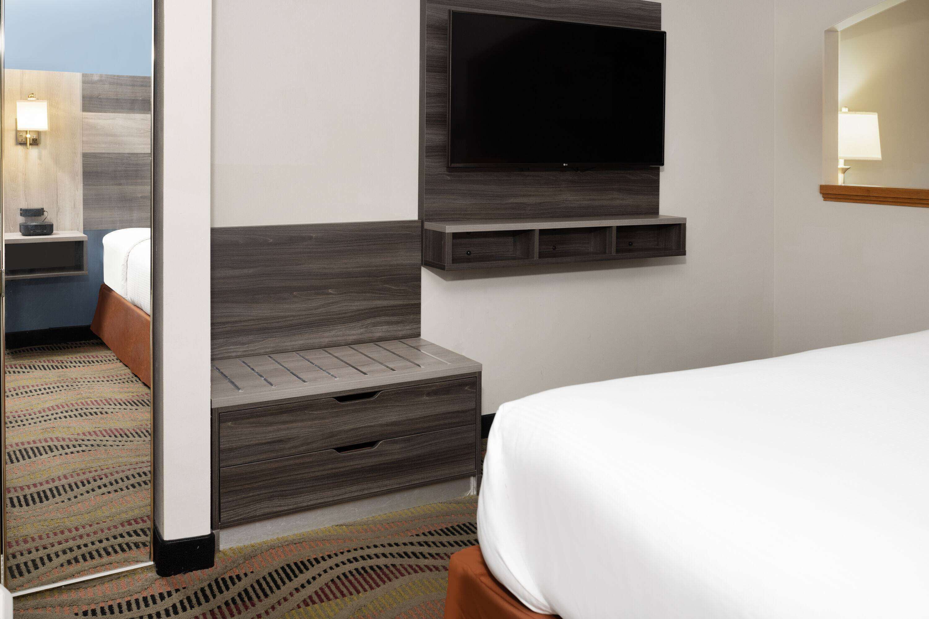 Habitación de hotel con televisor de pantalla plana montado en la pared, cabecera de madera, mesita de noche con lámpara y alfombra estampada.