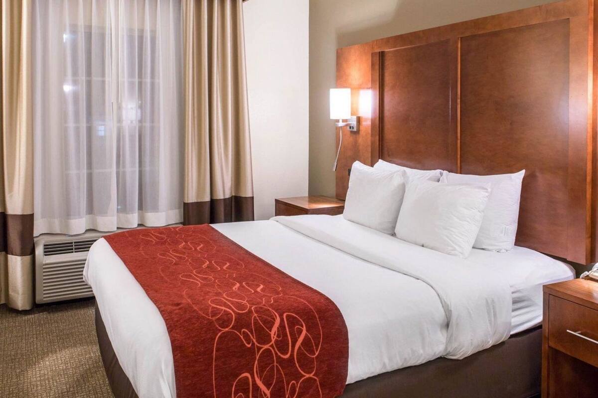 Habitación de hotel con una cama grande, una lámpara de noche, cabecera de madera y una ventana con cortinas.