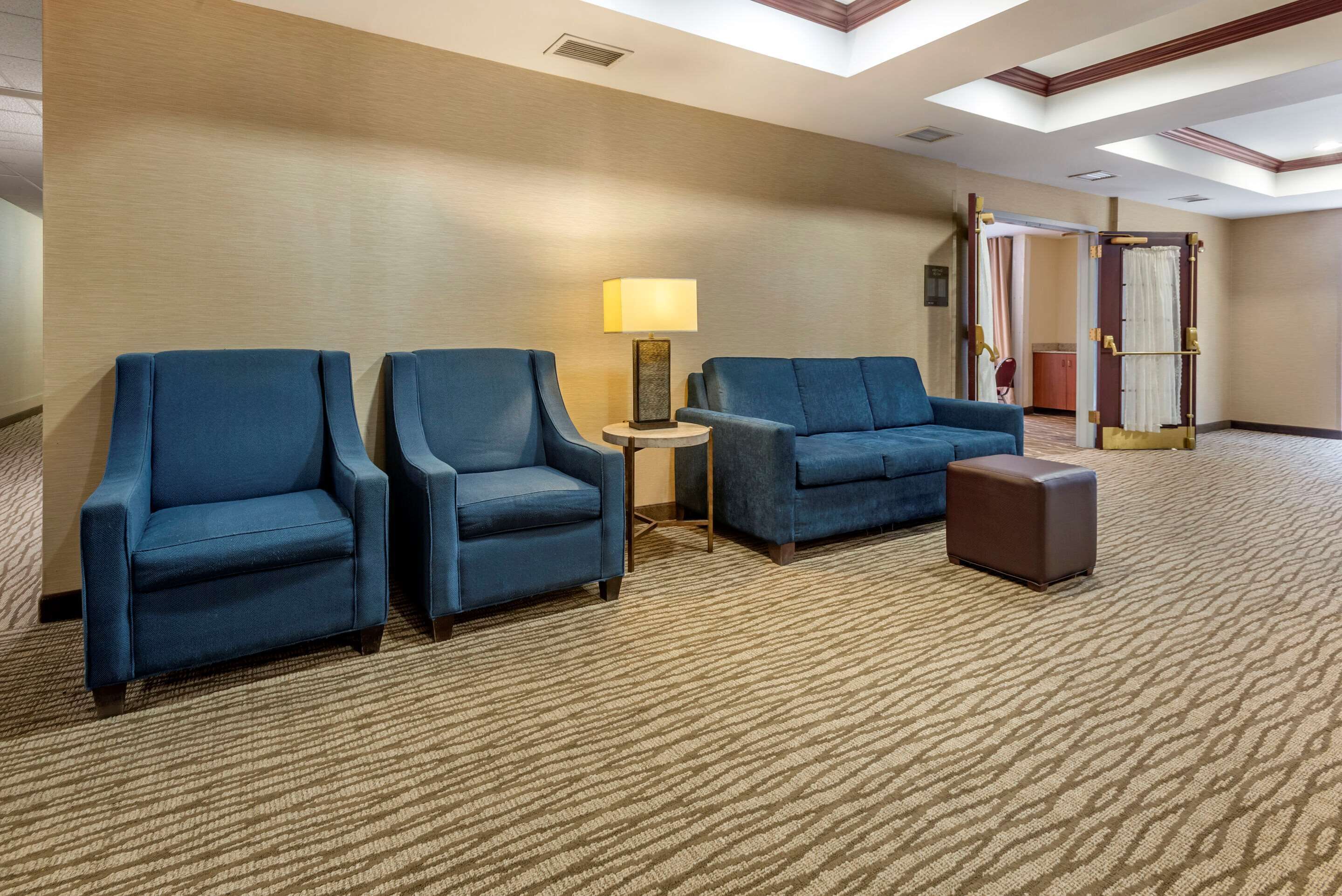 A hotel lobby with three blue armchairs, a small round table with a lamp, and a brown ottoman.
