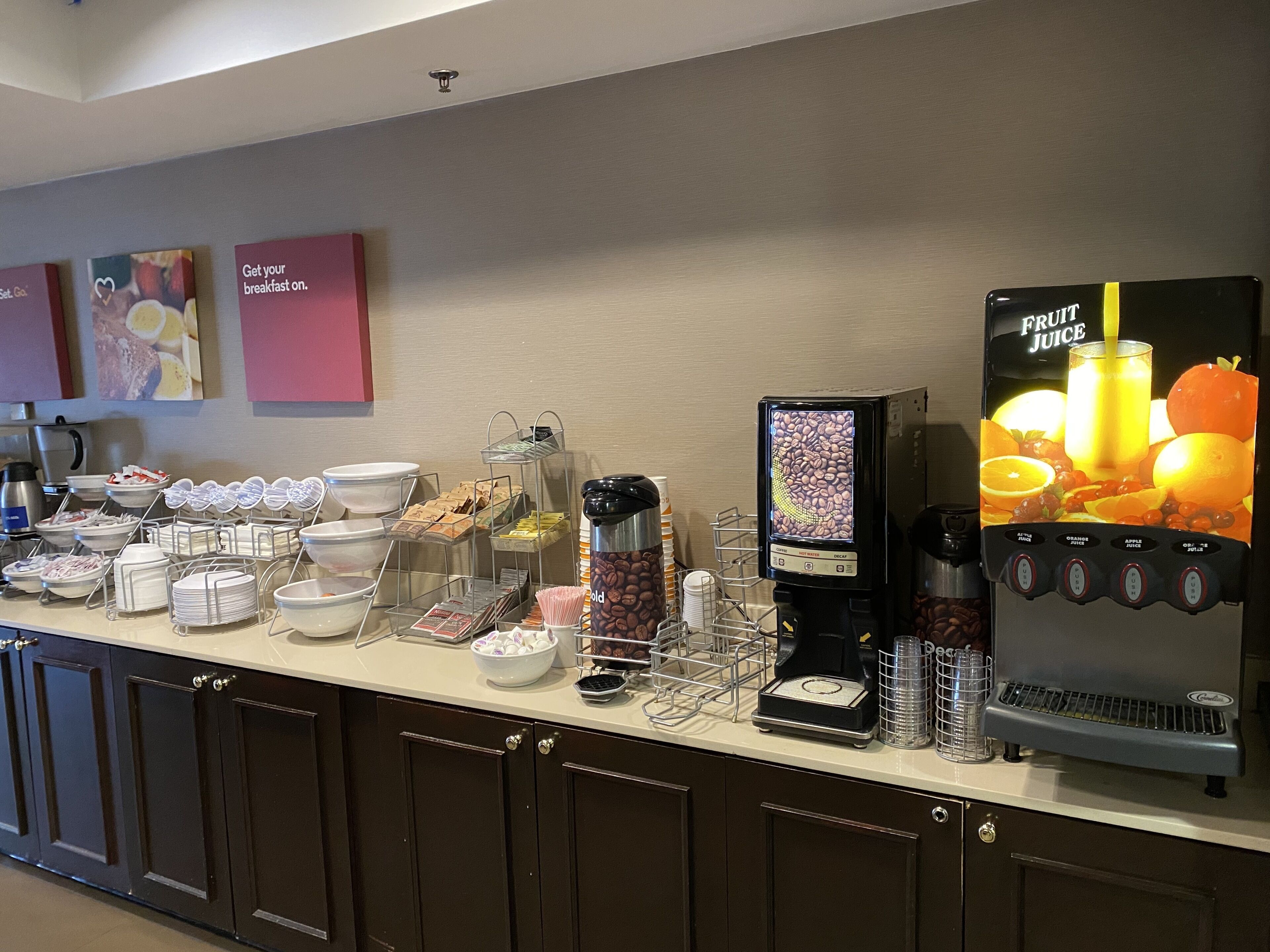 A breakfast bar with coffee machines, a fruit juice dispenser, and various food containers.