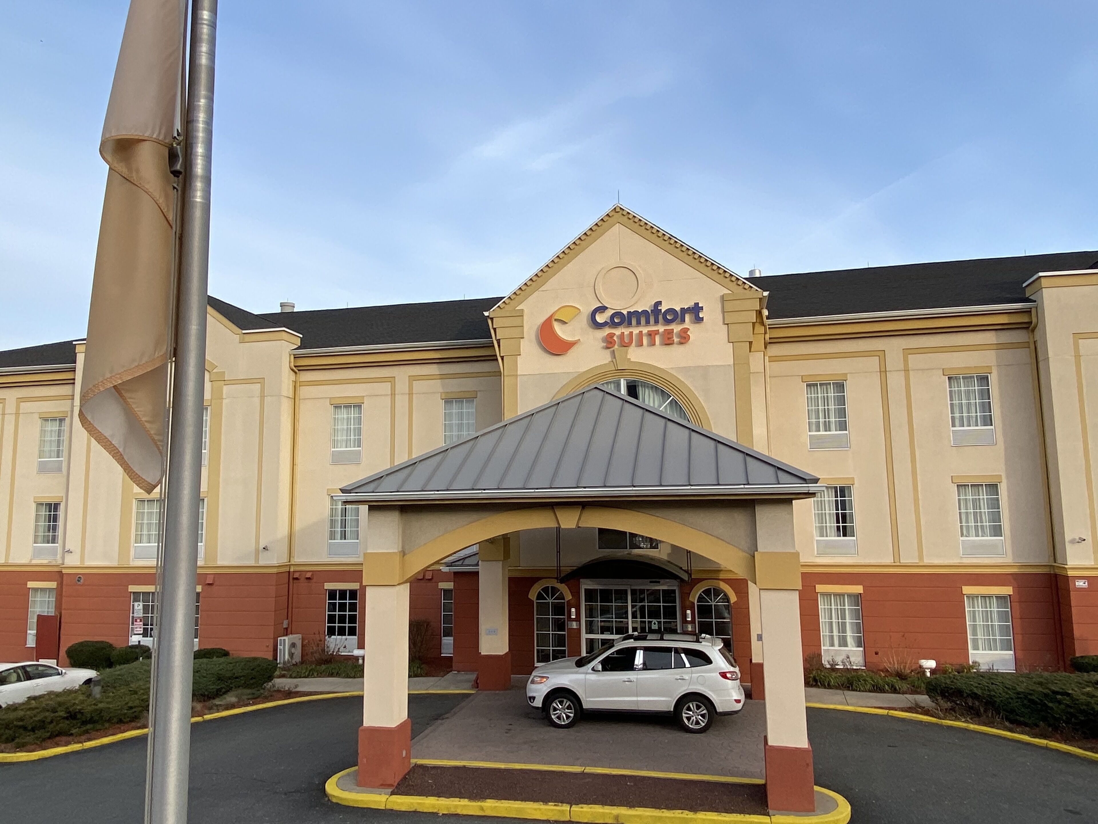 Un hotel Comfort Suites con un acceso cubierto, un coche blanco estacionado al frente y un asta de banderas a la izquierda.