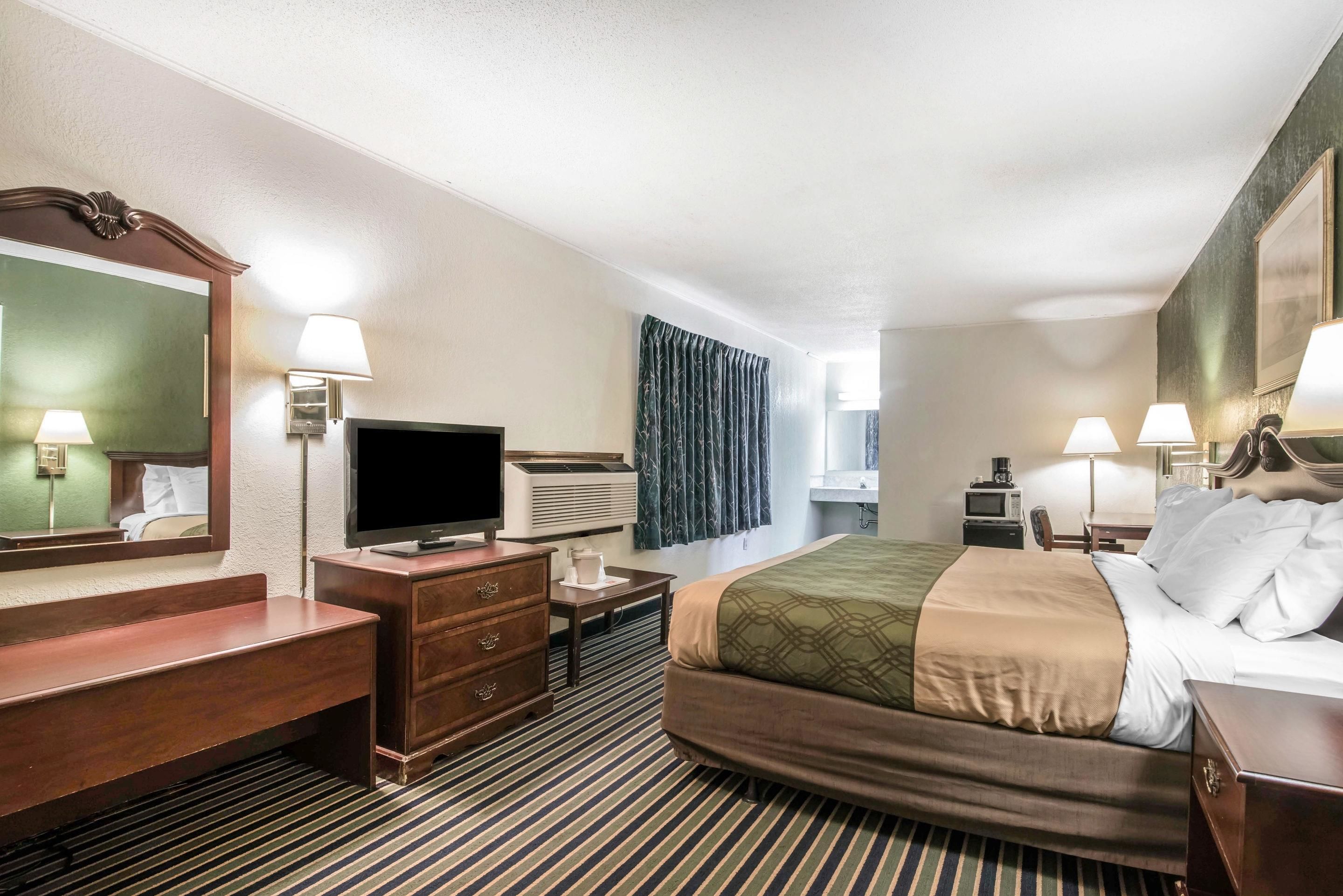 Quarto de hotel com cama, televisão, escrivaninha, espelho e janela com cortinas.