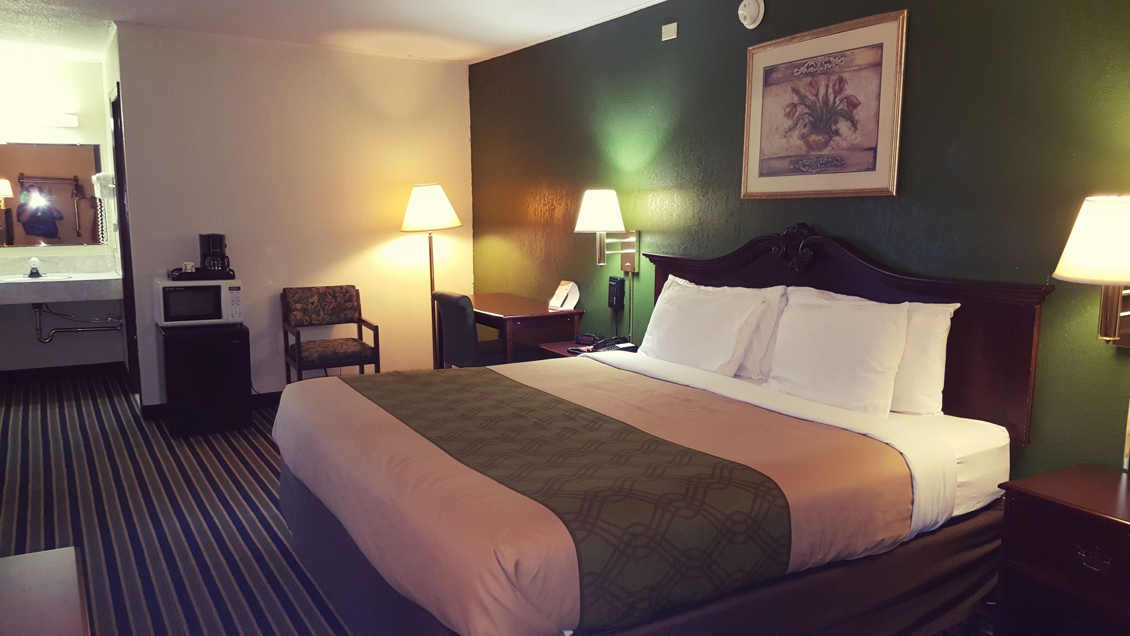 A hotel room with a bed, two chairs, a desk, a telephone, and a painting on the wall.