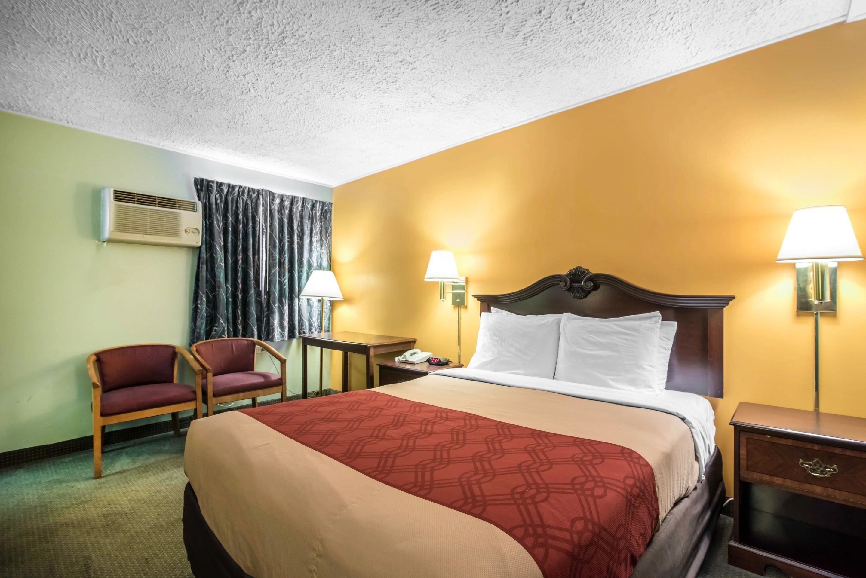 Una habitación de hotel con cama, dos sillas, una mesita de noche, lámpara y aire acondicionado.