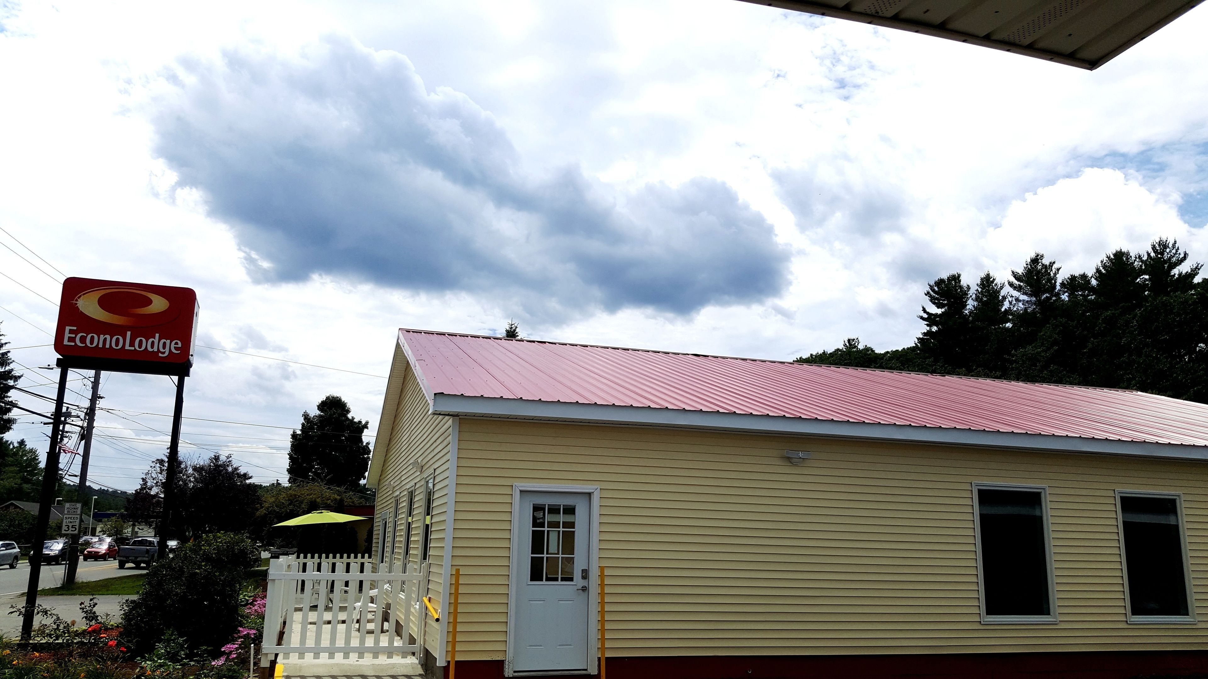 Un petit bâtiment d’un seul étage, avec un toit rouge et une porte blanche, entouré d’arbres et surplombé par un ciel nuageux.