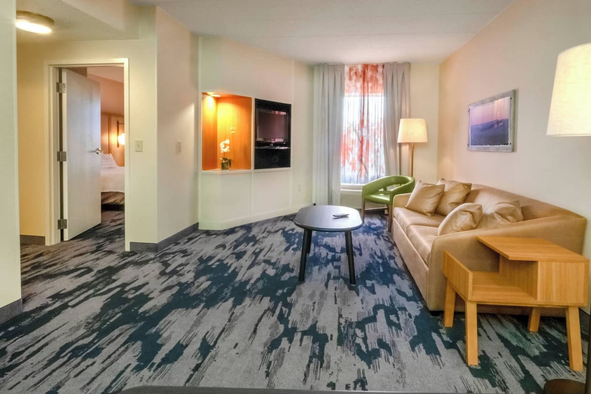 A hotel room with a beige sofa, a small wooden side table, a TV mounted on the wall, and a patterned carpet.