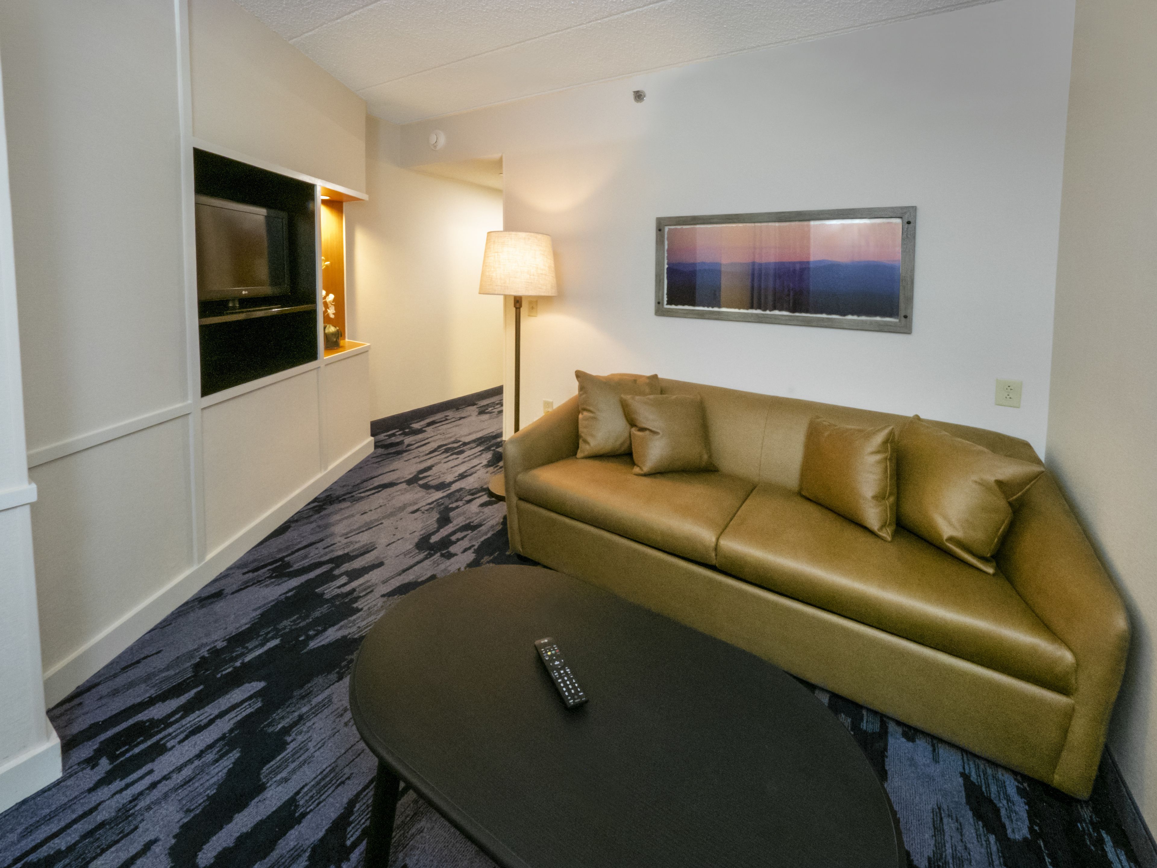 Ein Hotelzimmer mit einer Couch, einer Fernbedienung auf einem kleinen Tisch, einer Lampe und einem Fernseher auf einem Schrank.