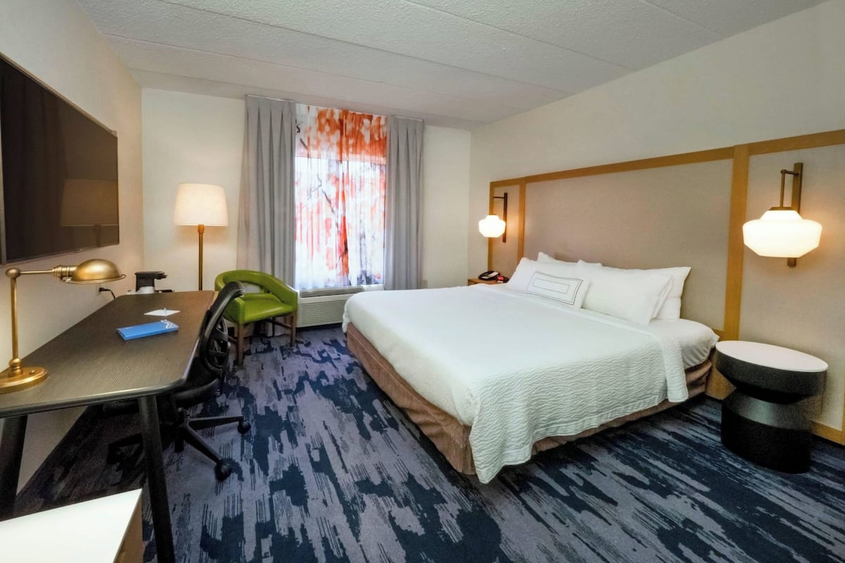 A hotel room with a bed, desk, chair, lamp, and a window with curtains.